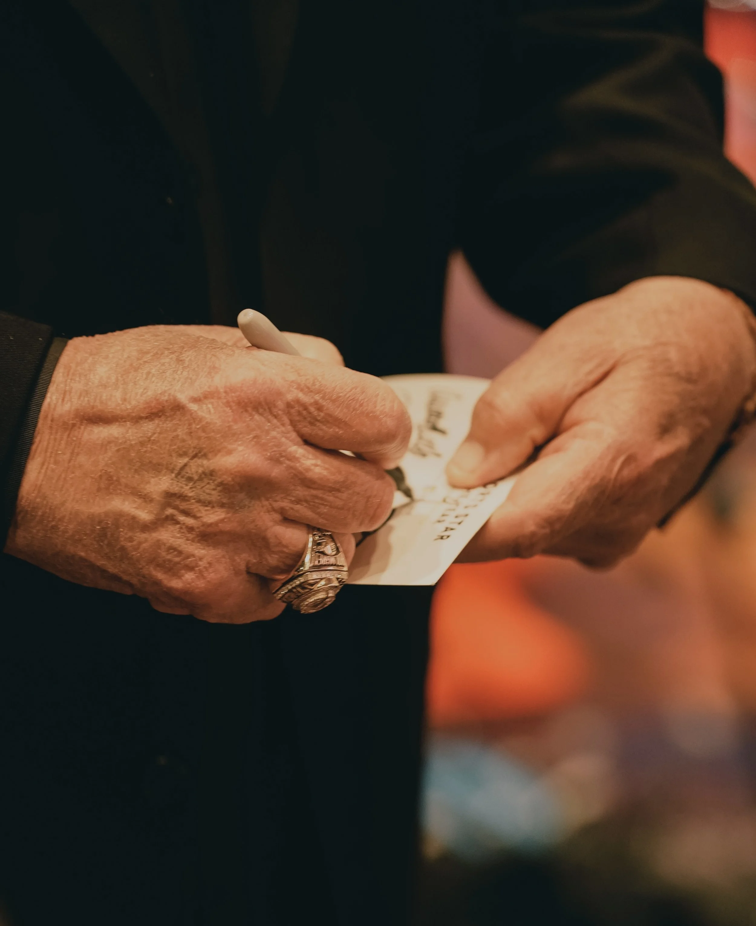 Person signing an item, possibly a piece of paper or a photograph, with a pen. The person's hands are visible, showing a large ring on one finger.