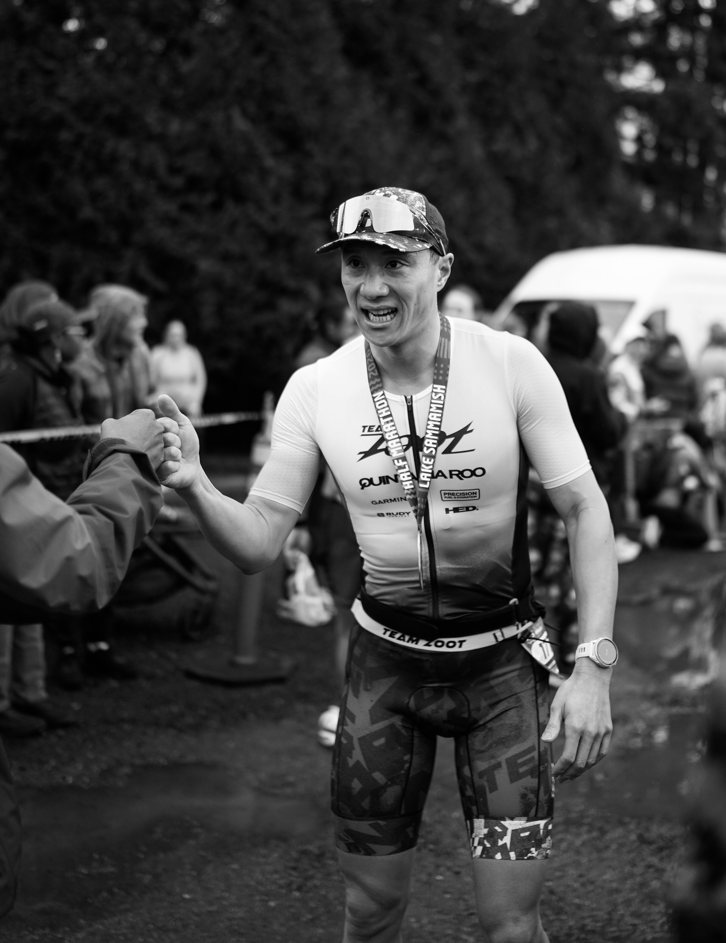 A male athlete in a finish line area, wearing a cap with sunglasses on top, medals around his neck, a race bib, and athletic clothing, engaging in a handshake.