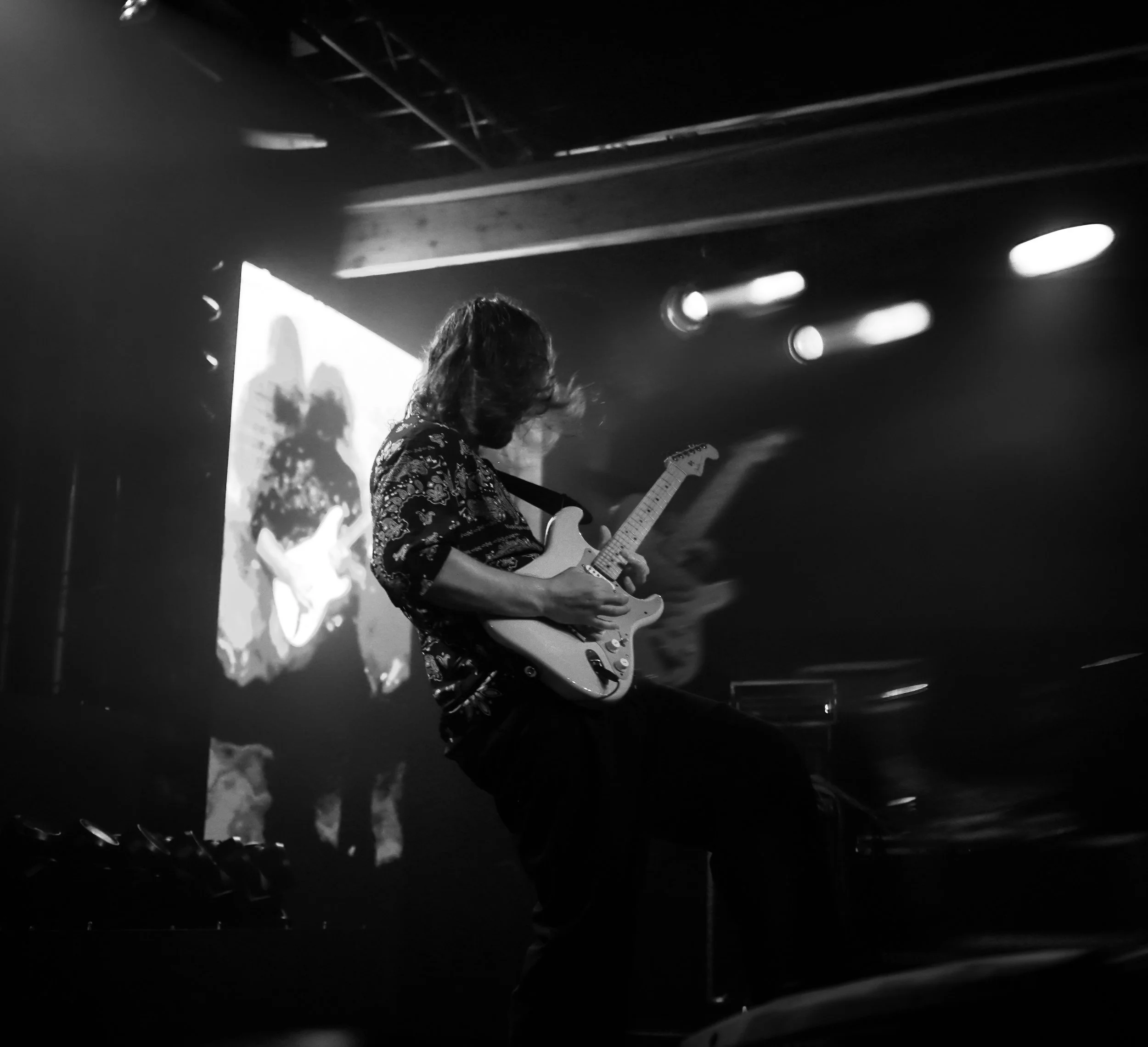 A musician with long hair playing an electric guitar on stage amidst concert lighting, black and white photograph.
