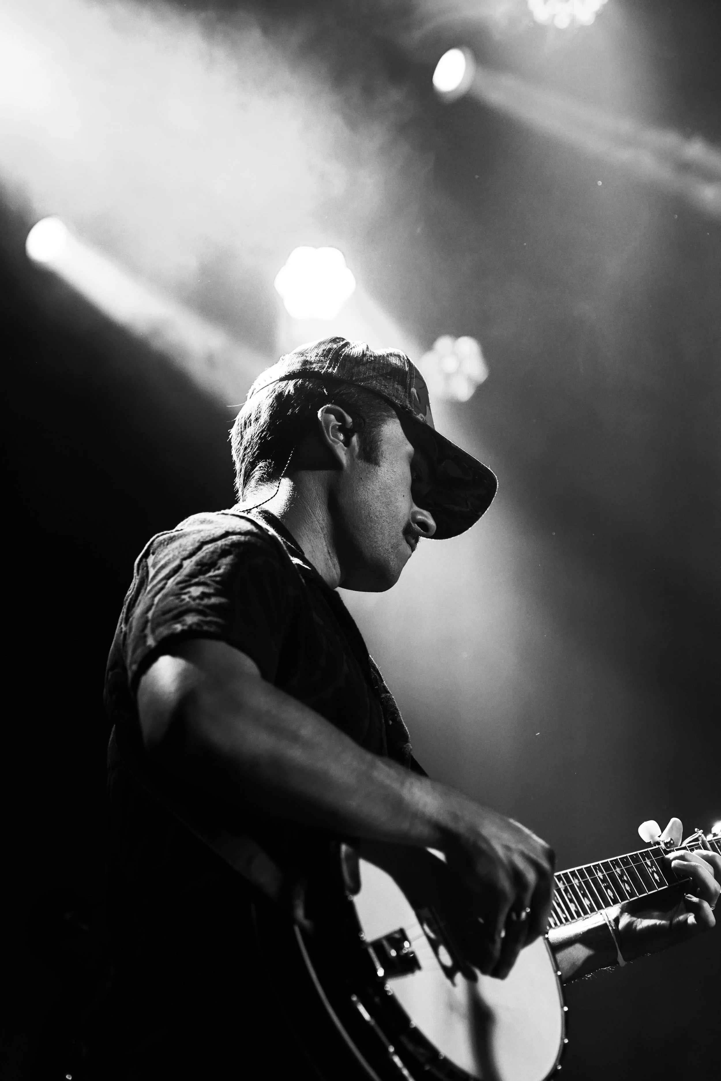 A young man wearing a cap and black clothing playing an electric guitar on stage, backlit with stage lights, black and white photo.