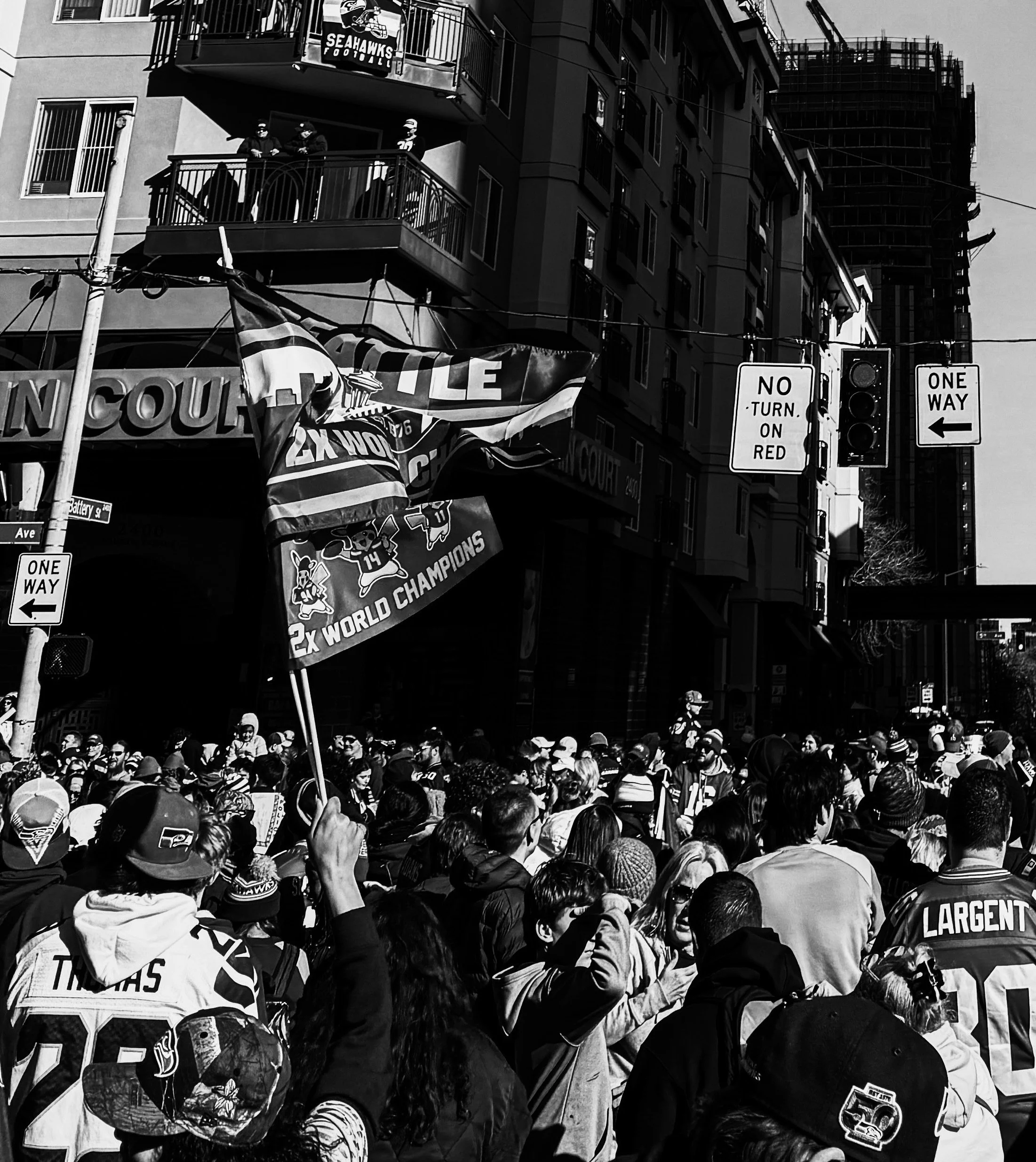 Crowd of Seattle Seahawks fans gathered on the street, some holding flags cheering for the team, with buildings and traffic lights in the background.