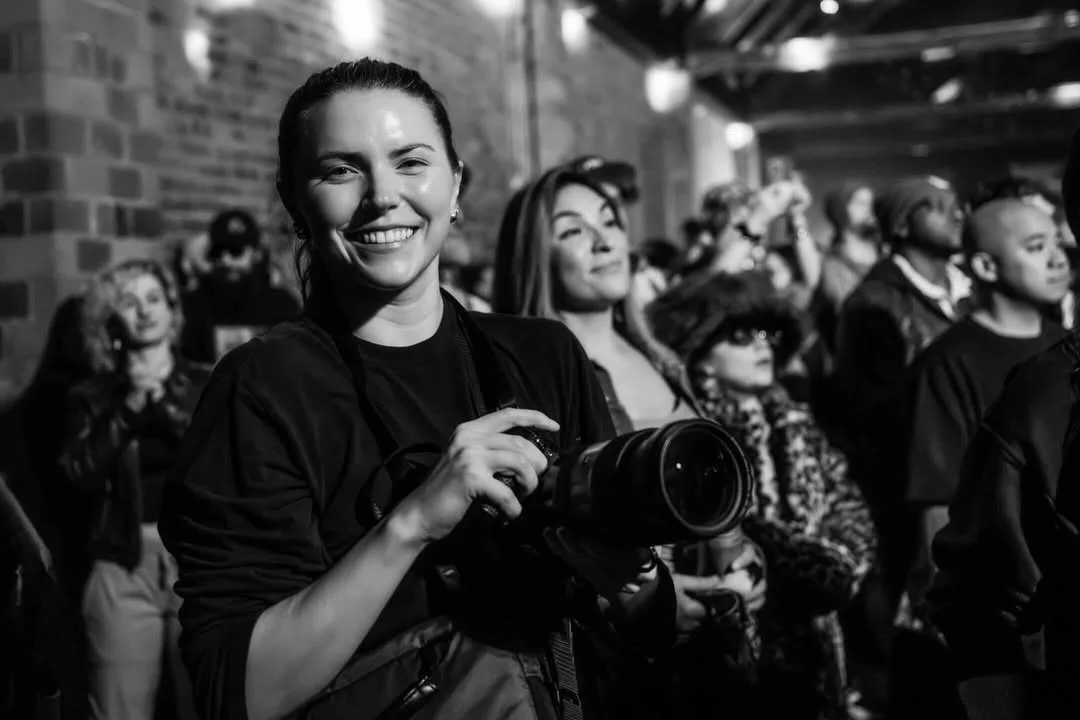 A smiling woman holding a professional camera at a crowded indoor event, with brick walls and other attendees in the background.