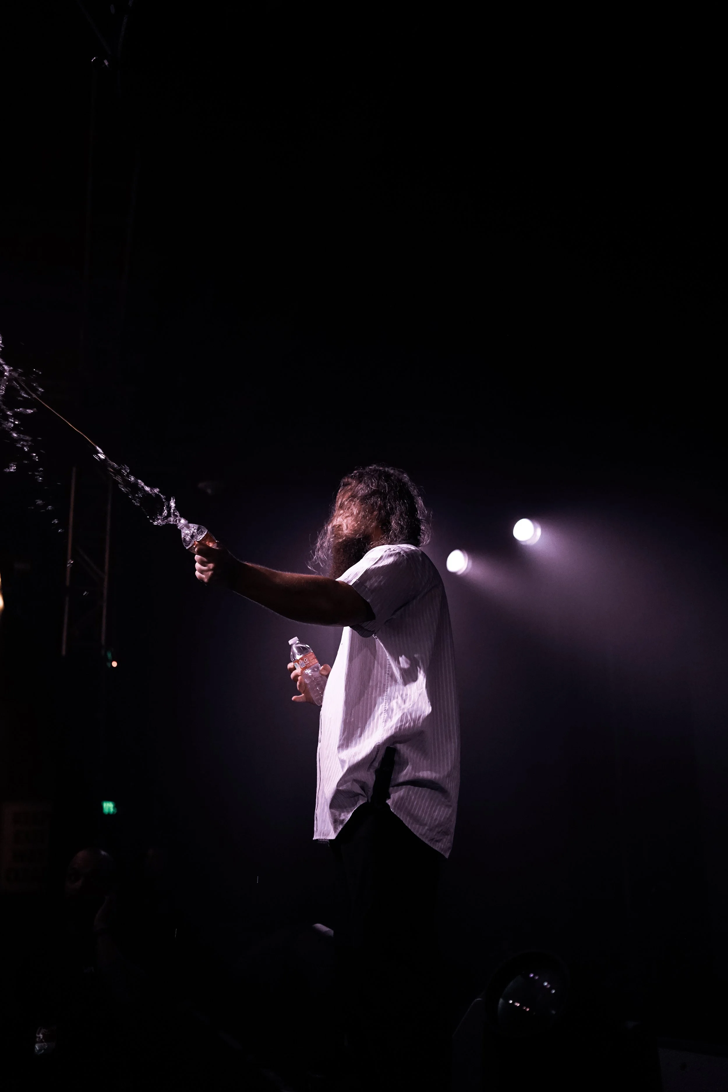 A man with long hair and a beard on stage, holding a water bottle and spraying water, with bright stage lights in the background.