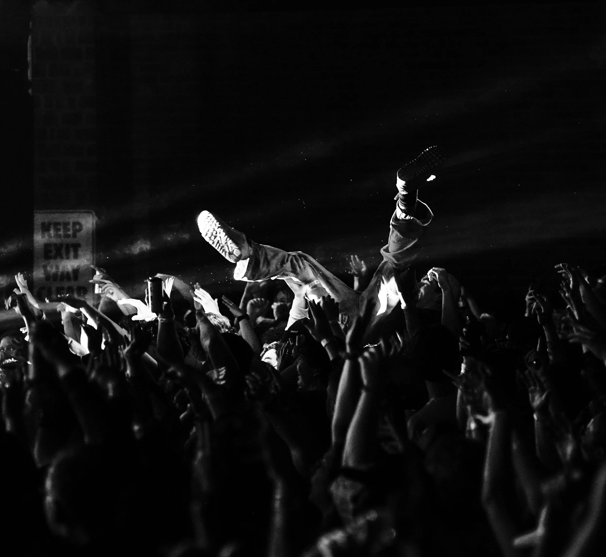 A crowd at a concert with one person being lifted above others, creating a silhouette against the dark background, as the audience reaches out their hands.