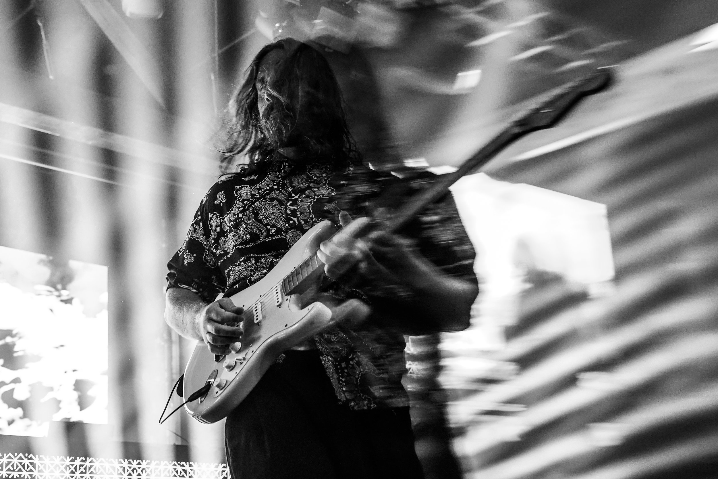 A musician with long hair playing an electric guitar on stage during a concert, with lights and motion blur in the background.