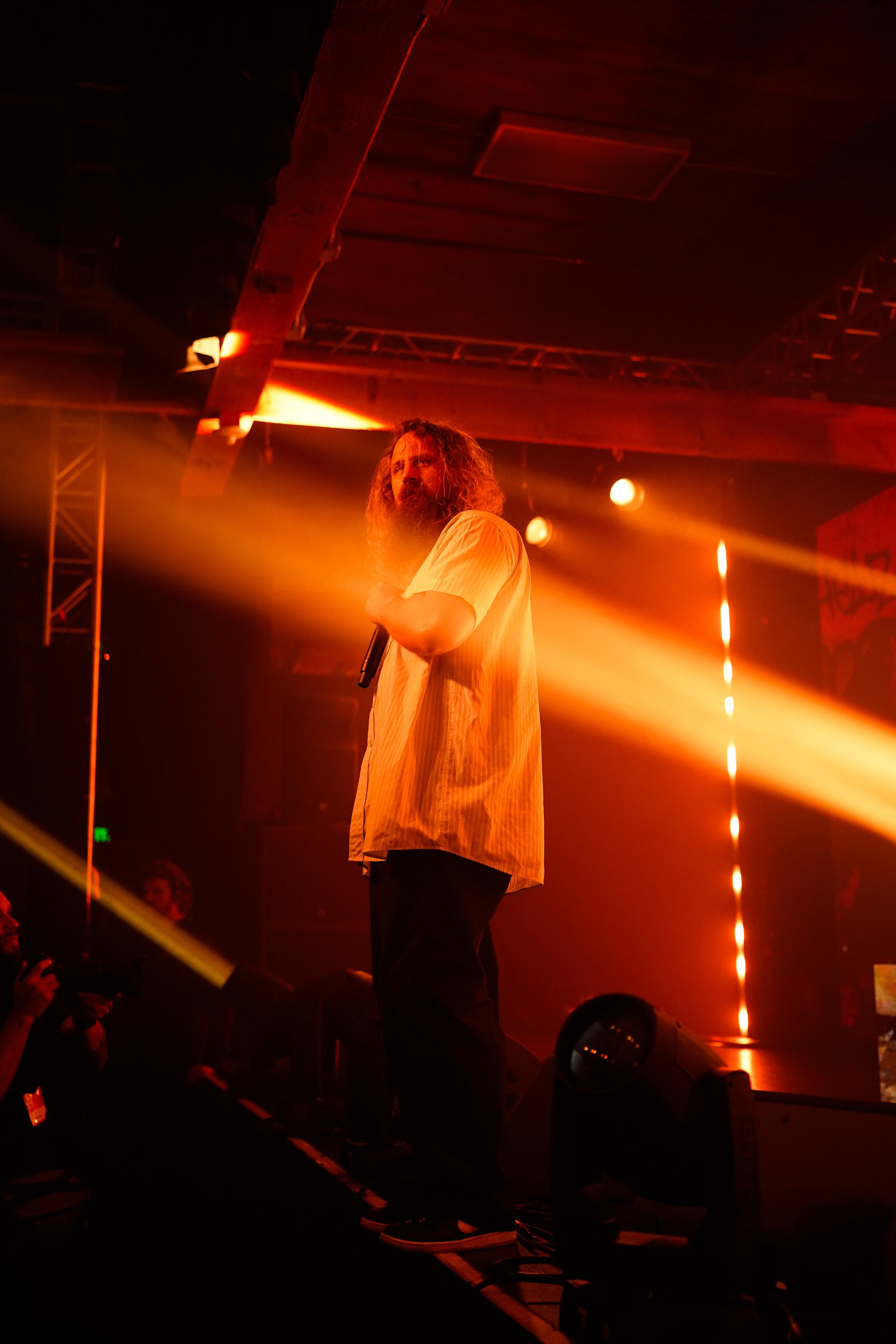 A man with long, curly hair and a beard holds a microphone on stage illuminated by warm orange spotlights in a dark venue.