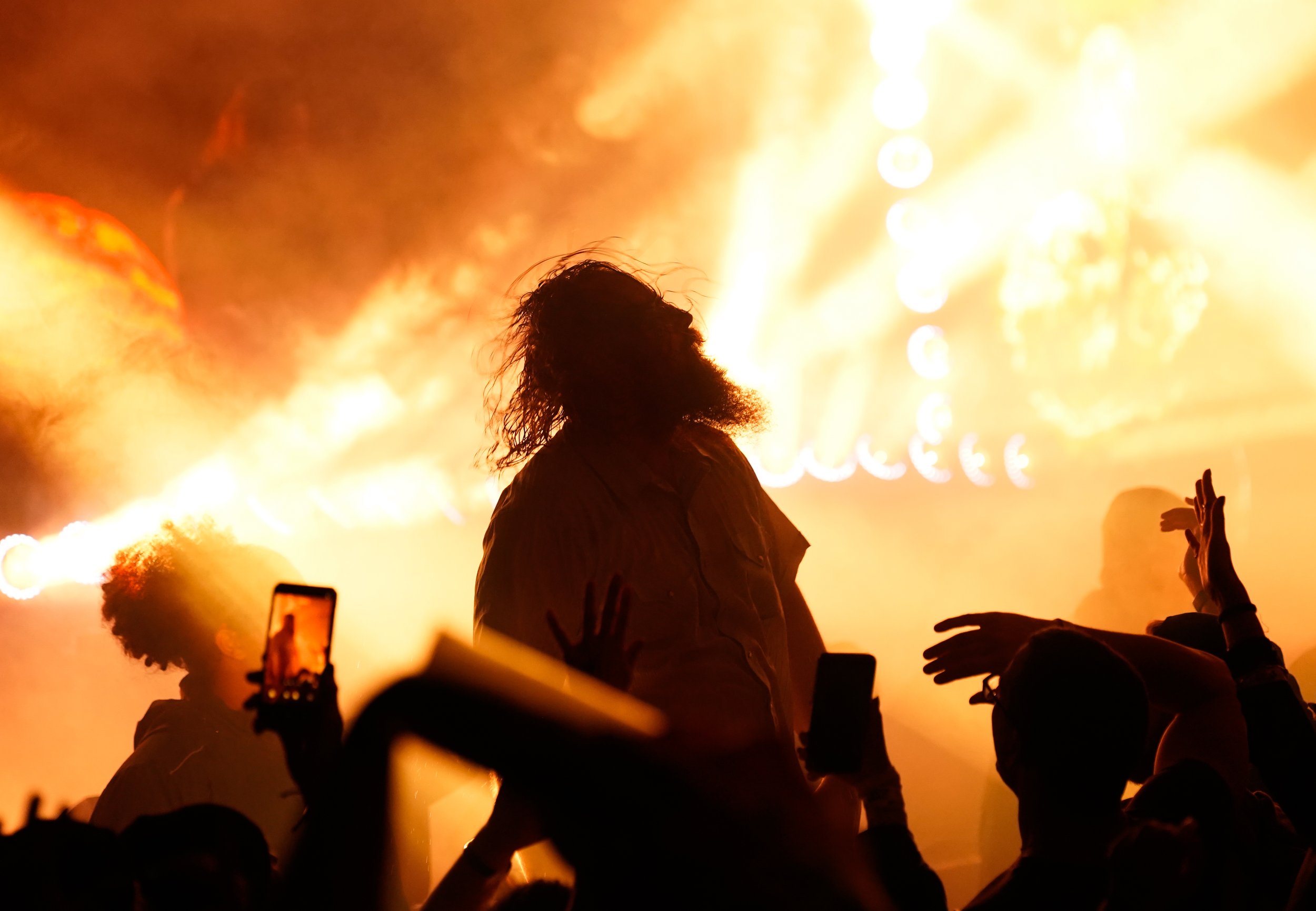 Silhouette of a person with curly hair performing on stage at a concert, with bright orange and yellow stage lights and smoke in the background, while audience members reach out and record with phones.