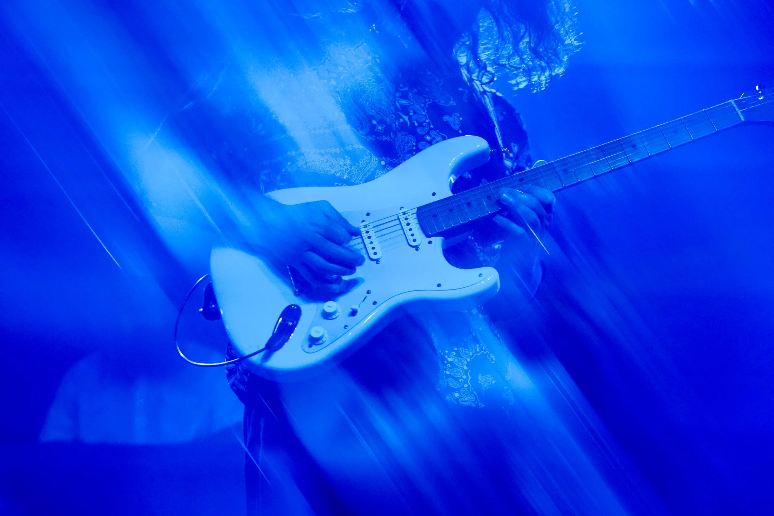 Person playing a white electric guitar on stage illuminated with blue light, with a patterned shirt and long hair.