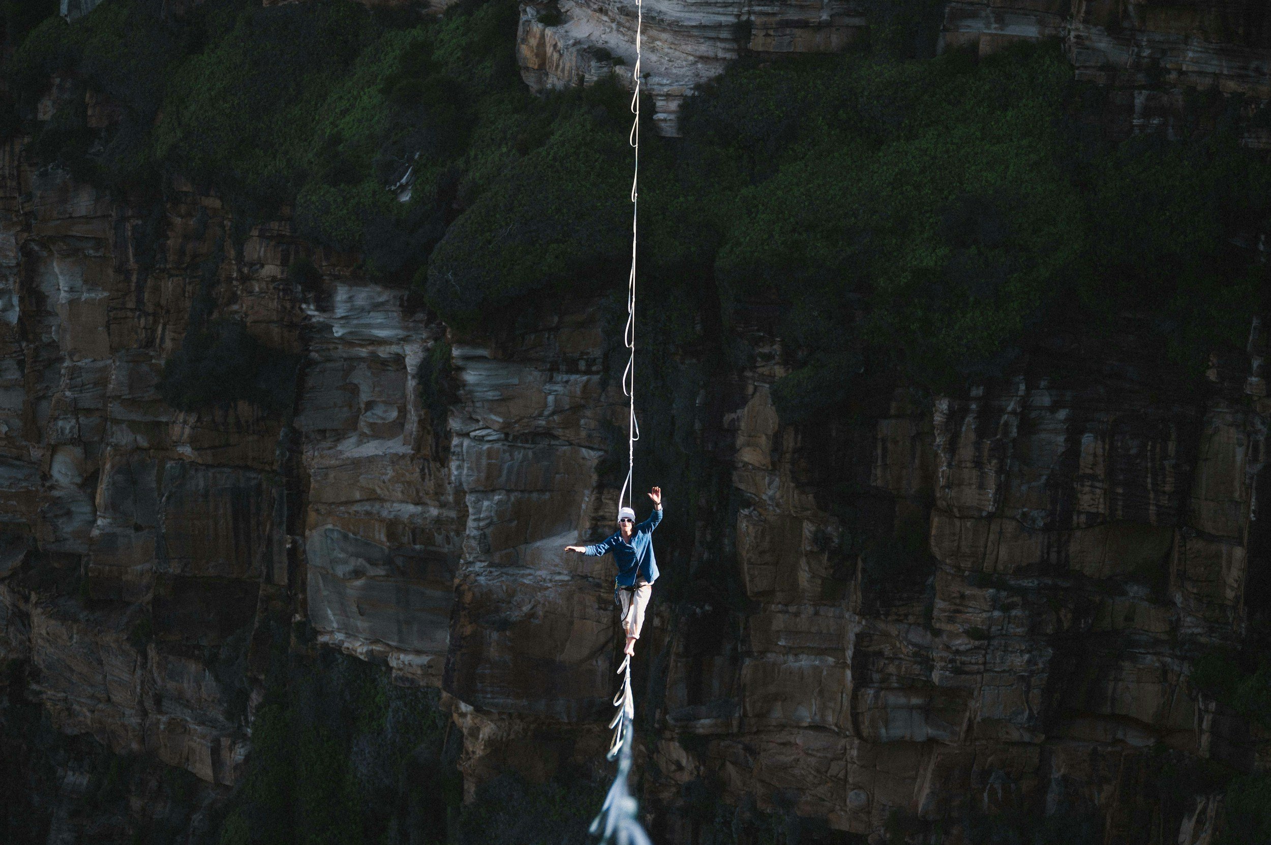A person in a blue jacket and white hat is walking on a tightrope stretched high above a rocky canyon with green vegetation.