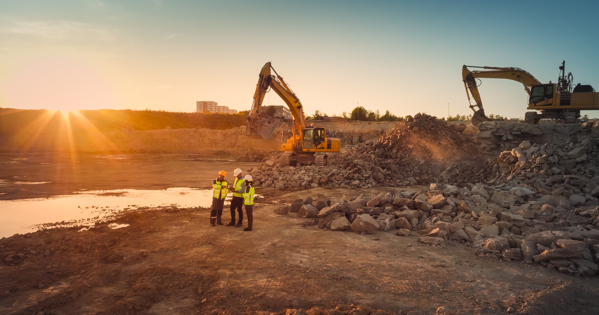 Construction site during sunset with two excavators and three workers wearing safety vests and helmets, standing near a body of water.