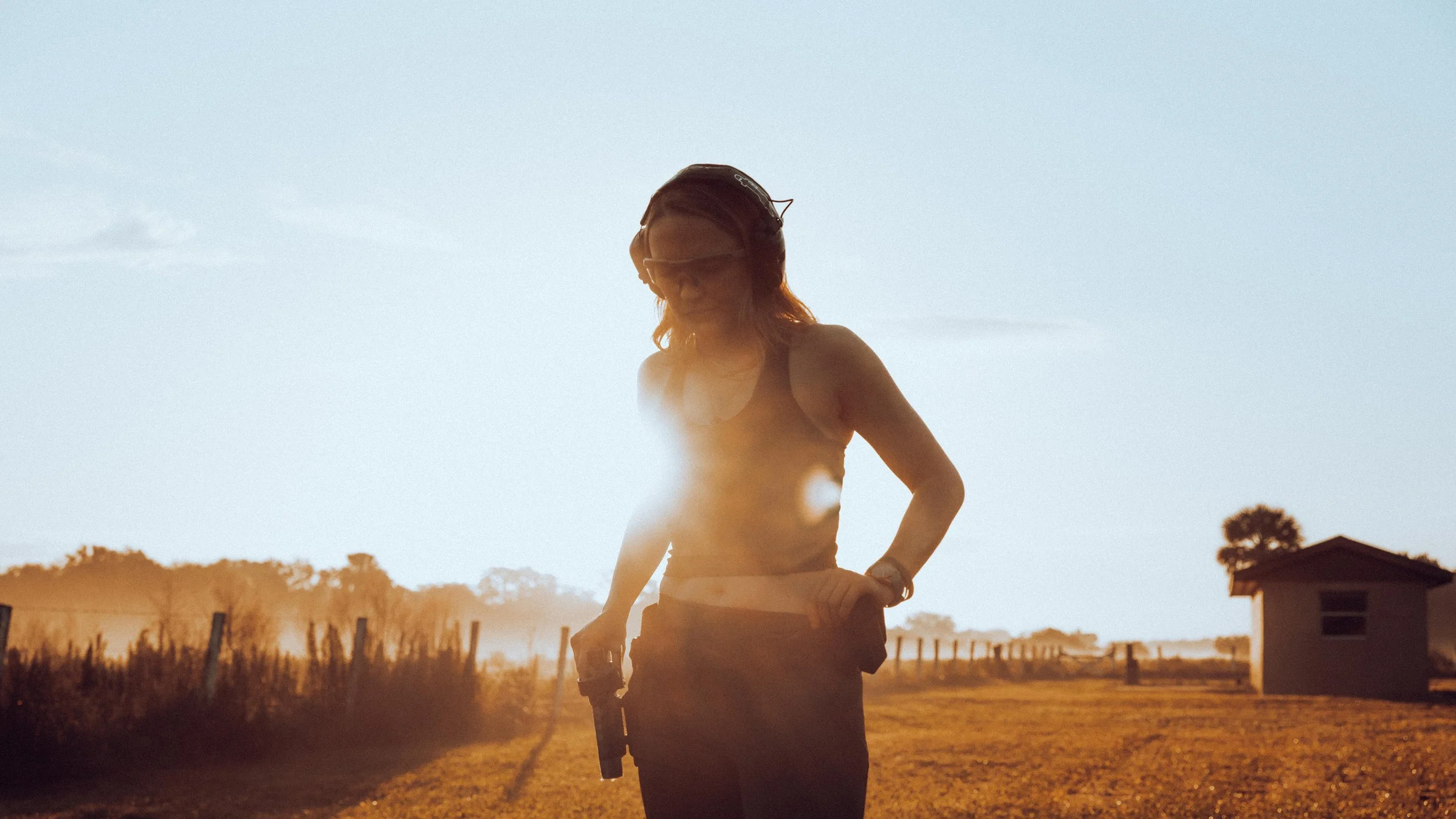 A woman outdoors during sunset wearing sunglasses and a helmet, holding a golf club, with trees and a house in the background.