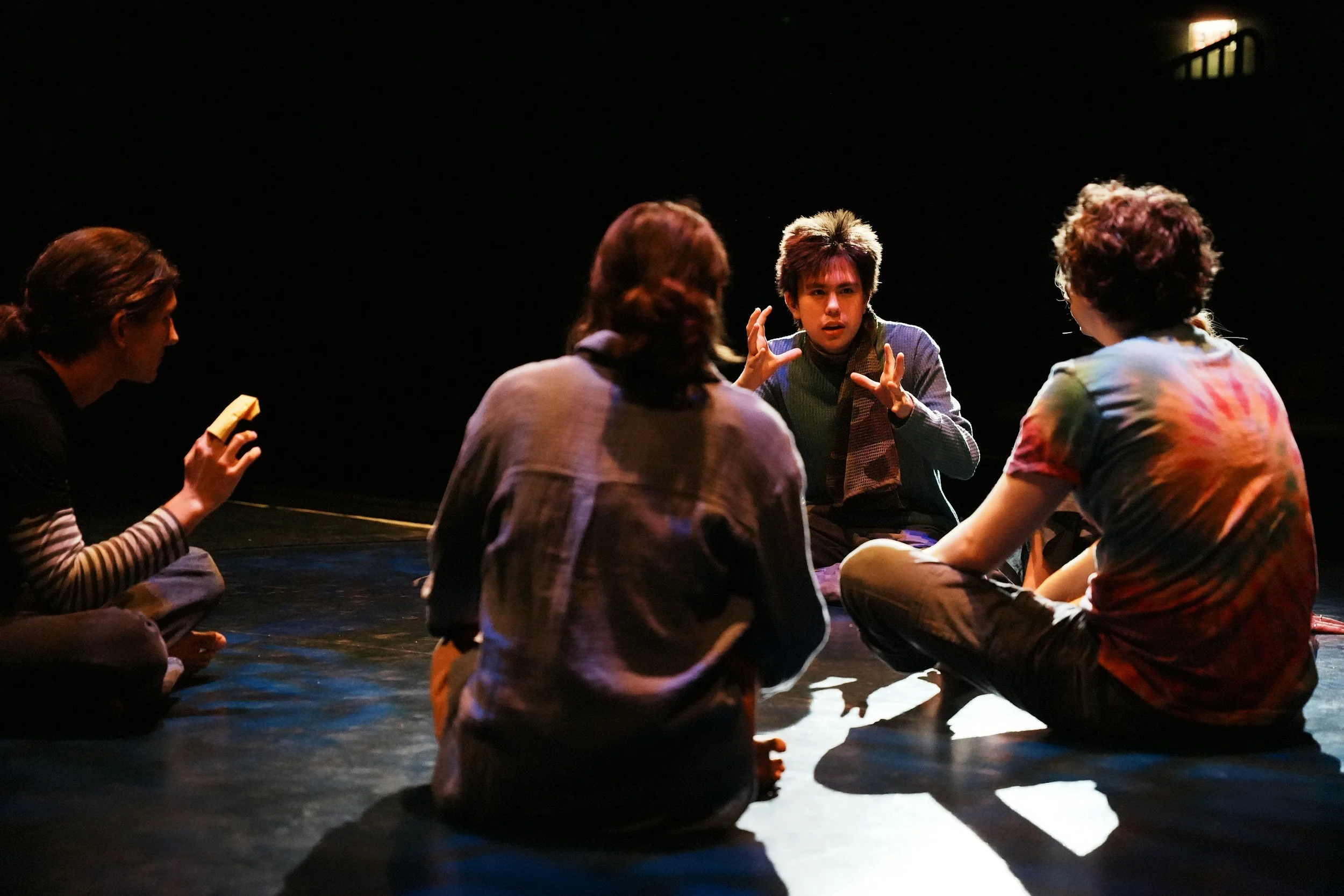 A group of five people sitting on the floor in a circle having a discussion or conversation in a dimly lit room.
