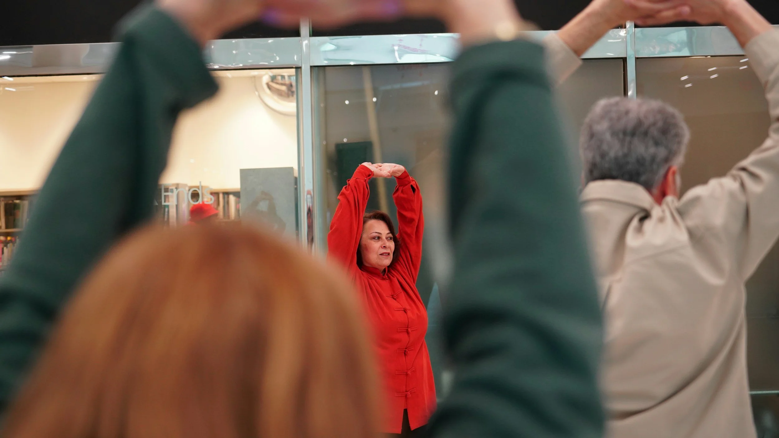 A woman in a red jacket stretching or exercising with her arms raised above her head, seen through a crowd of people in a library.