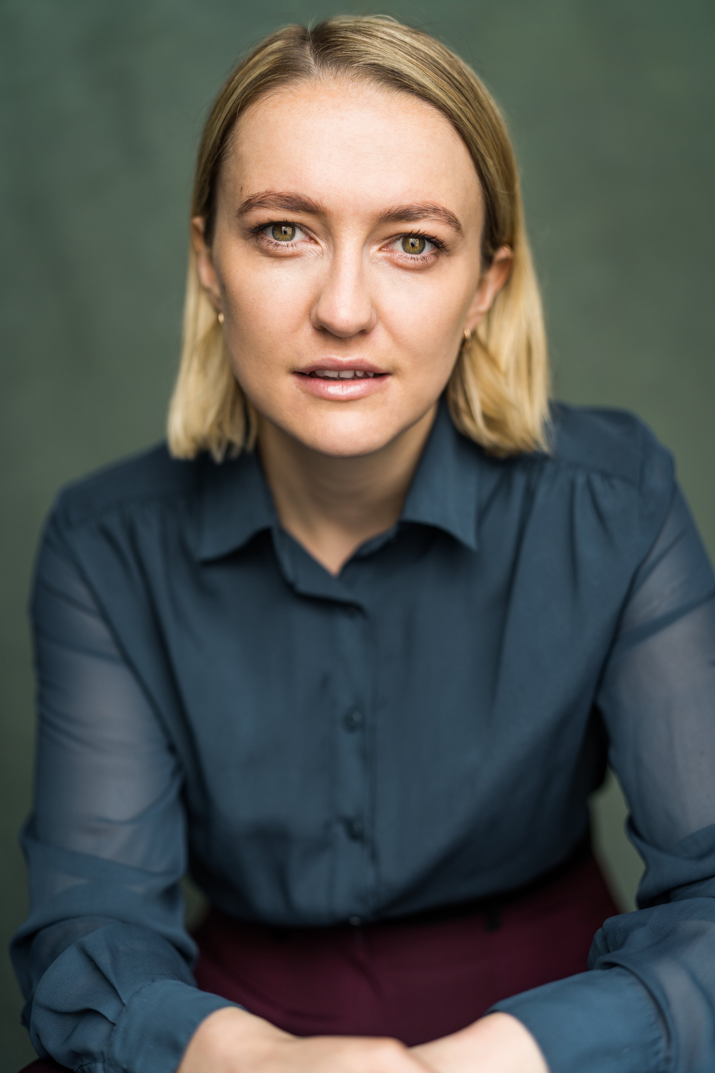 Close-up of a woman with blonde hair, green eyes, wearing a dark blue shirt, sitting with her arms crossed, looking directly at the camera.