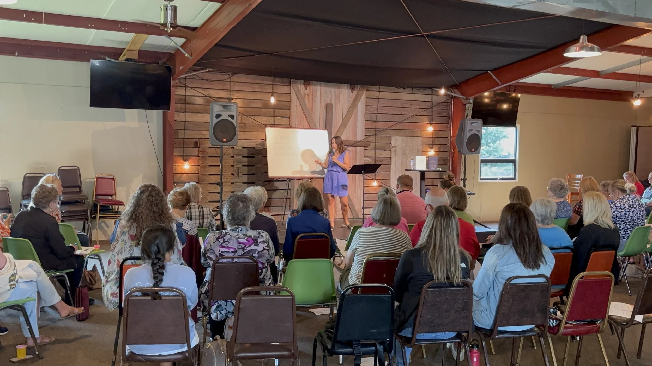 Christian speaker and author Kayla Craig in a blue dress is giving a presentation to an audience seated on chairs in a rustic indoor space with wood-paneled walls and warm lighting at Okoboji Bible Conference.