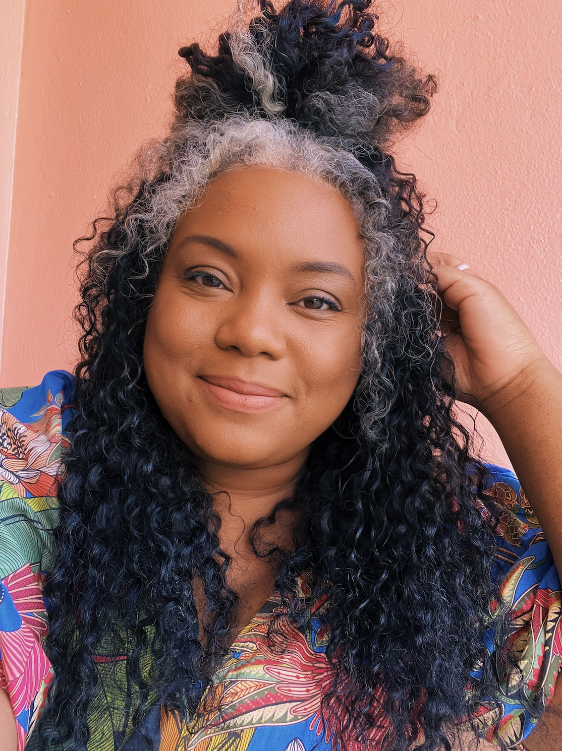 Headshot of Morgan Nichols, author and poet. Woman with curly black and gray hair, smiling, wearing a colorful floral shirt, against a pink wall.
