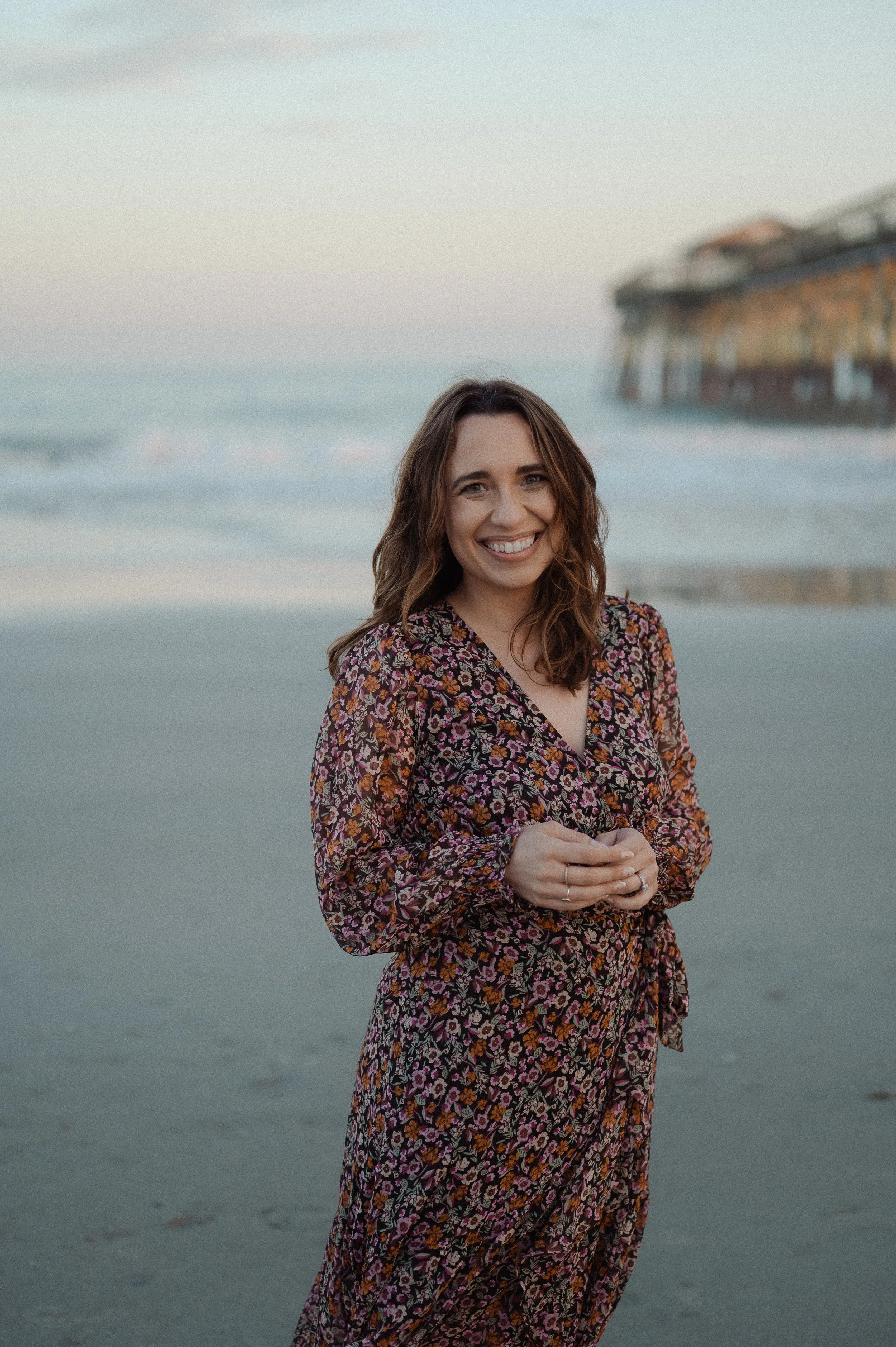 Bestelling contemplative Christian author Kayla Craig with wavy brown hair and a floral dress smiling on the beach with a pier in the background.