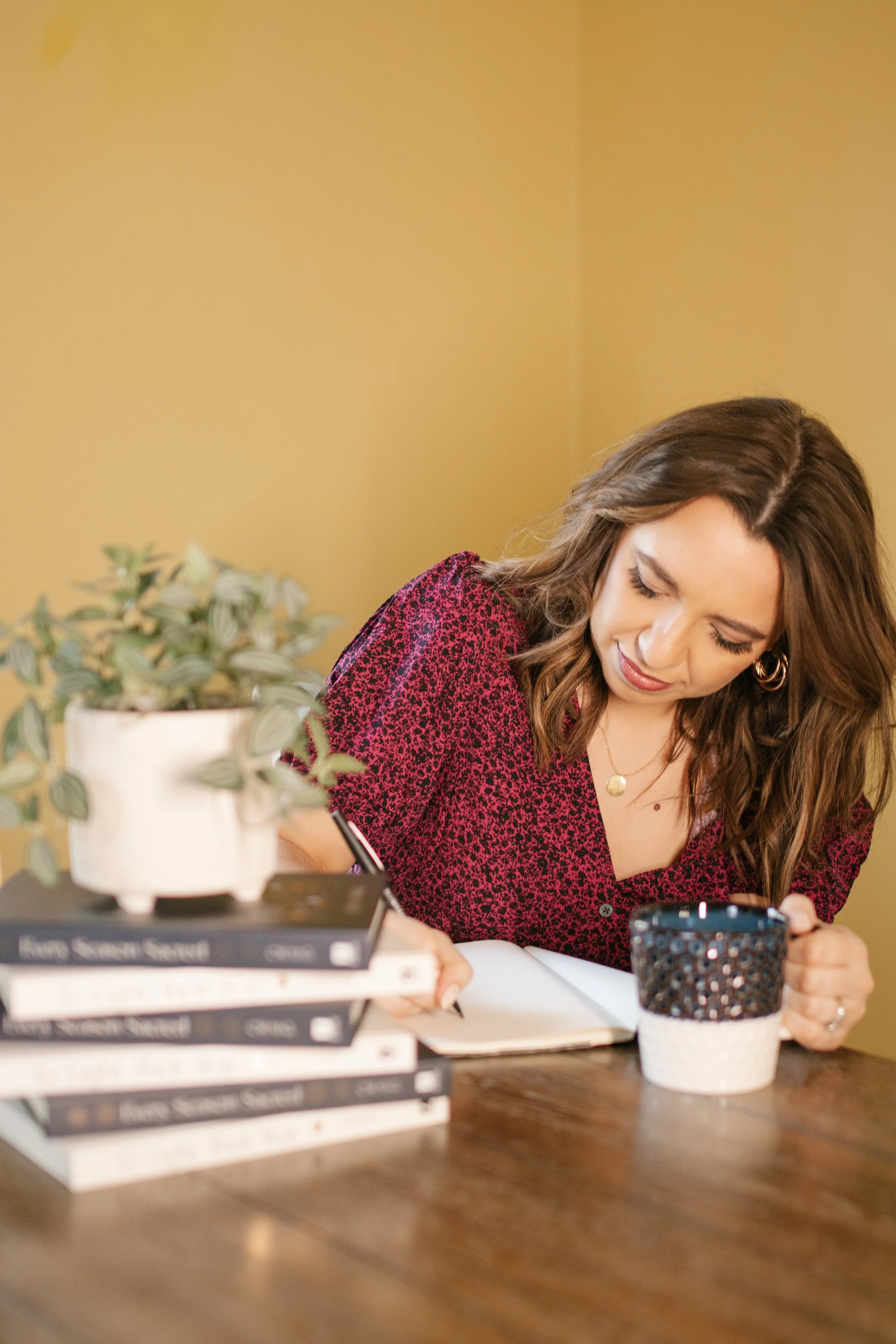Author Kayla Craig writing in her journal. A stack of her beautiful hardcover books Every Season Sacred and To Light Their Way are stacked on the table next to her.