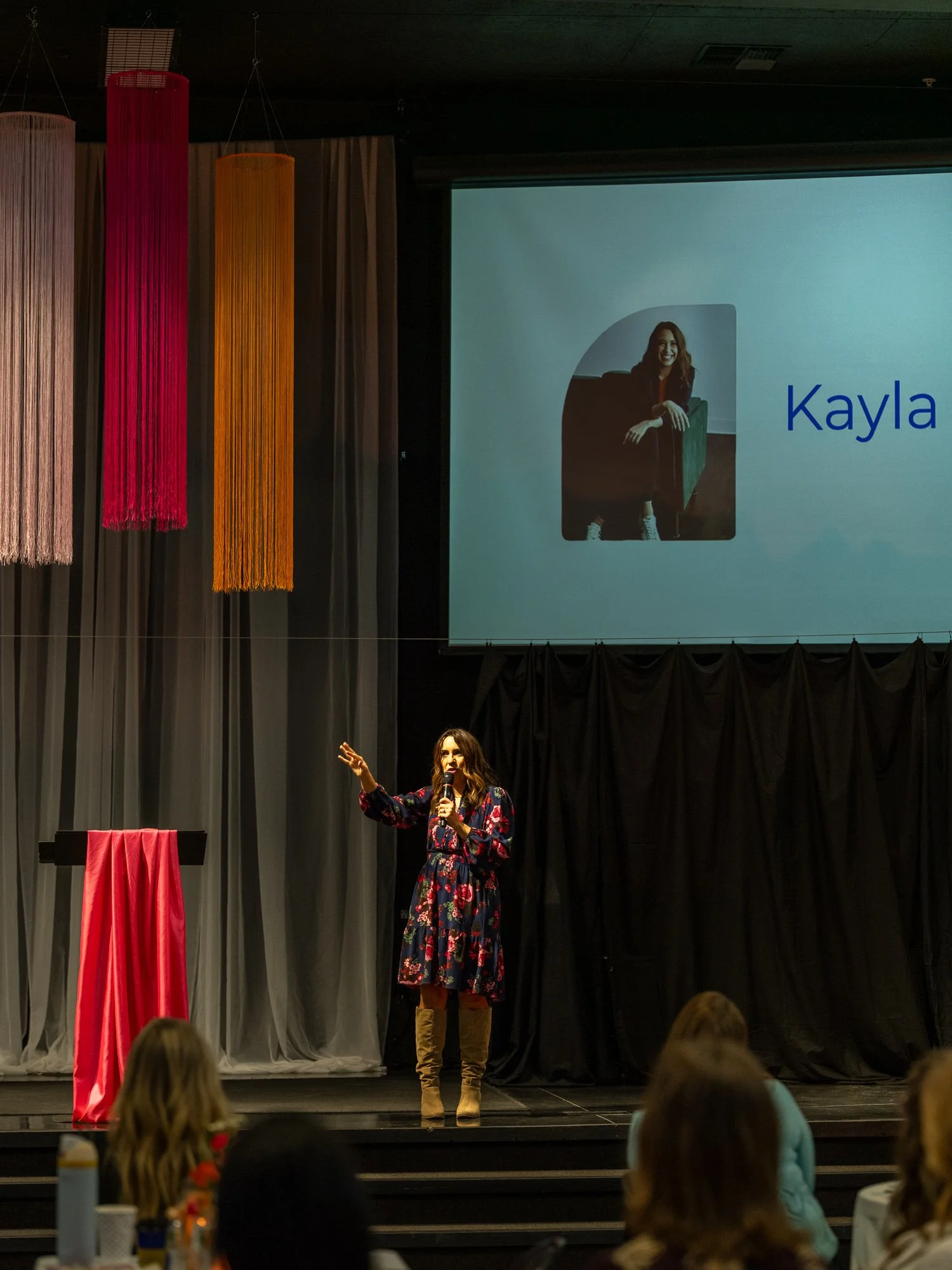 Speaker Kayla Craig in a floral dress and beige boots is speaking into a microphone on a stage with a black curtain backdrop. Behind her is a large screen displaying a woman with the name Kayla.