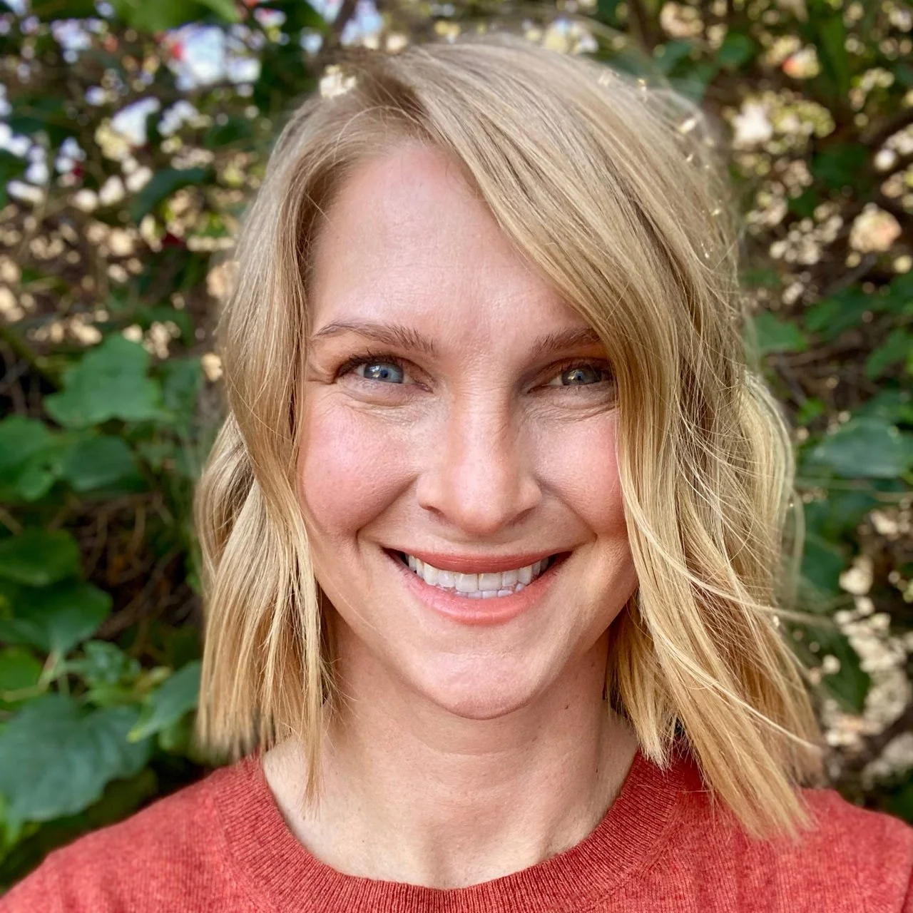 Headshot of bestselling author Meredith Miller. Smiling woman with blonde hair and blue eyes, wearing a red top, standing outdoors in front of green foliage.