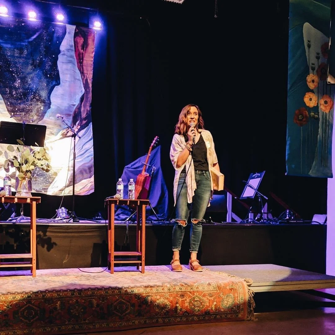 Author Kayla Craig standing on a stage holding a microphone, with a book in her other hand, in a setting that appears to be an event or performance space. There is a backdrop with artistic images and flowers on the side.