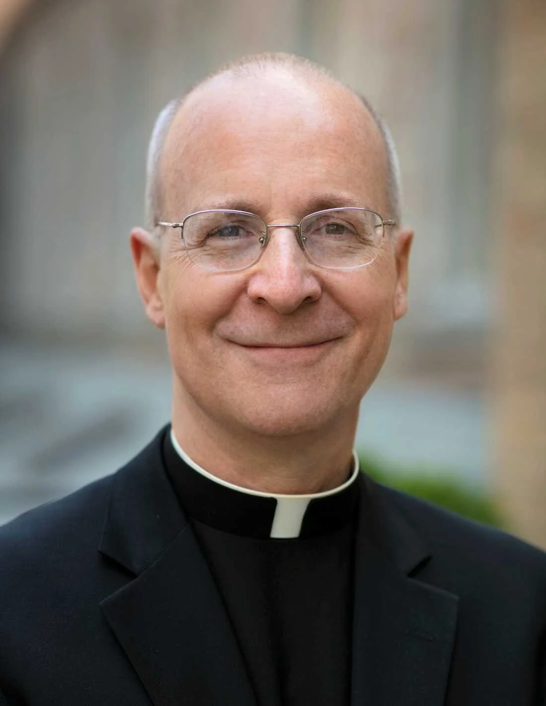Headshot of bestselling author Father James Martin, smiling man wearing glasses and a clerical collar, dressed in a black suit.