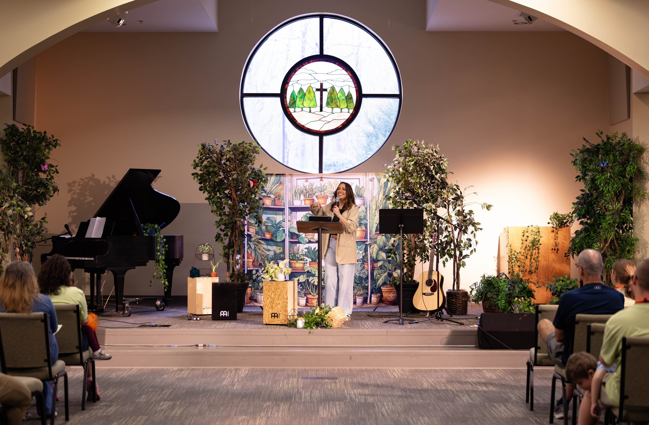 Author Kayla Craig speaking or performing on a stage decorated with plants and a colorful backdrop of potted plants. There is a stained-glass window behind her featuring trees, a cross, and a landscape. A church audience is seated in front.