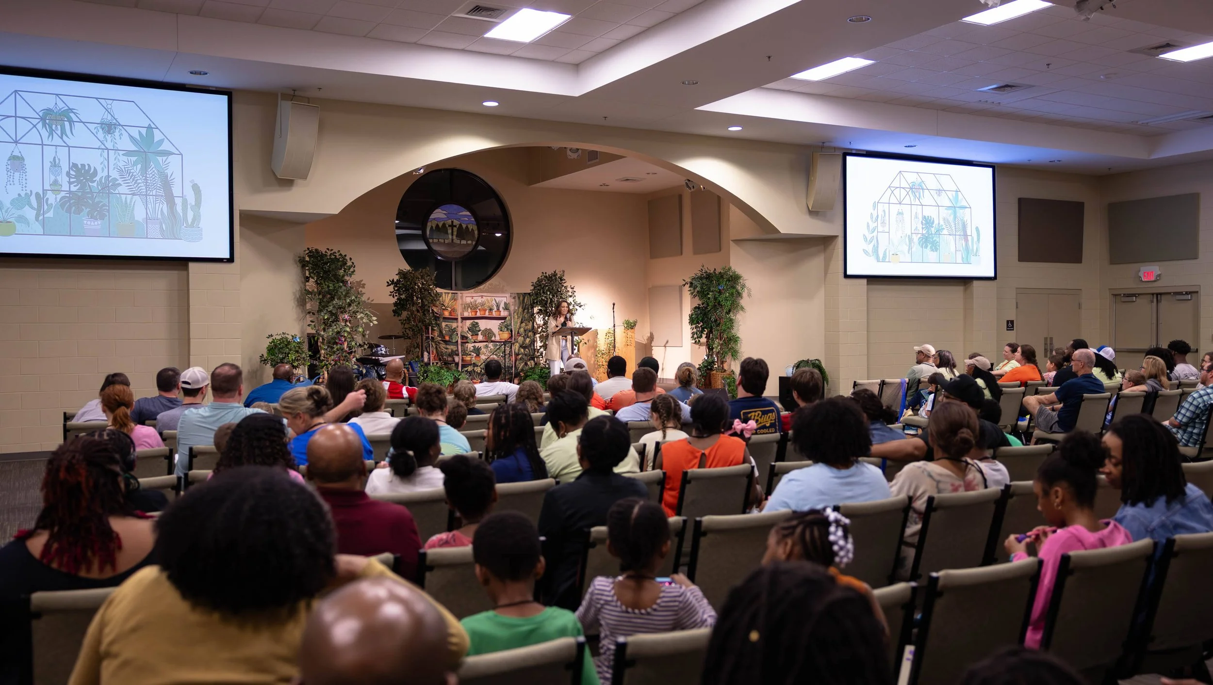 A large indoor auditorium filled with seated audience members watching speaker Kayla Craig peaking at a podium on stage. The stage has plants and a bookshelf with houseplants, and large screens on either side display the same decorative image.