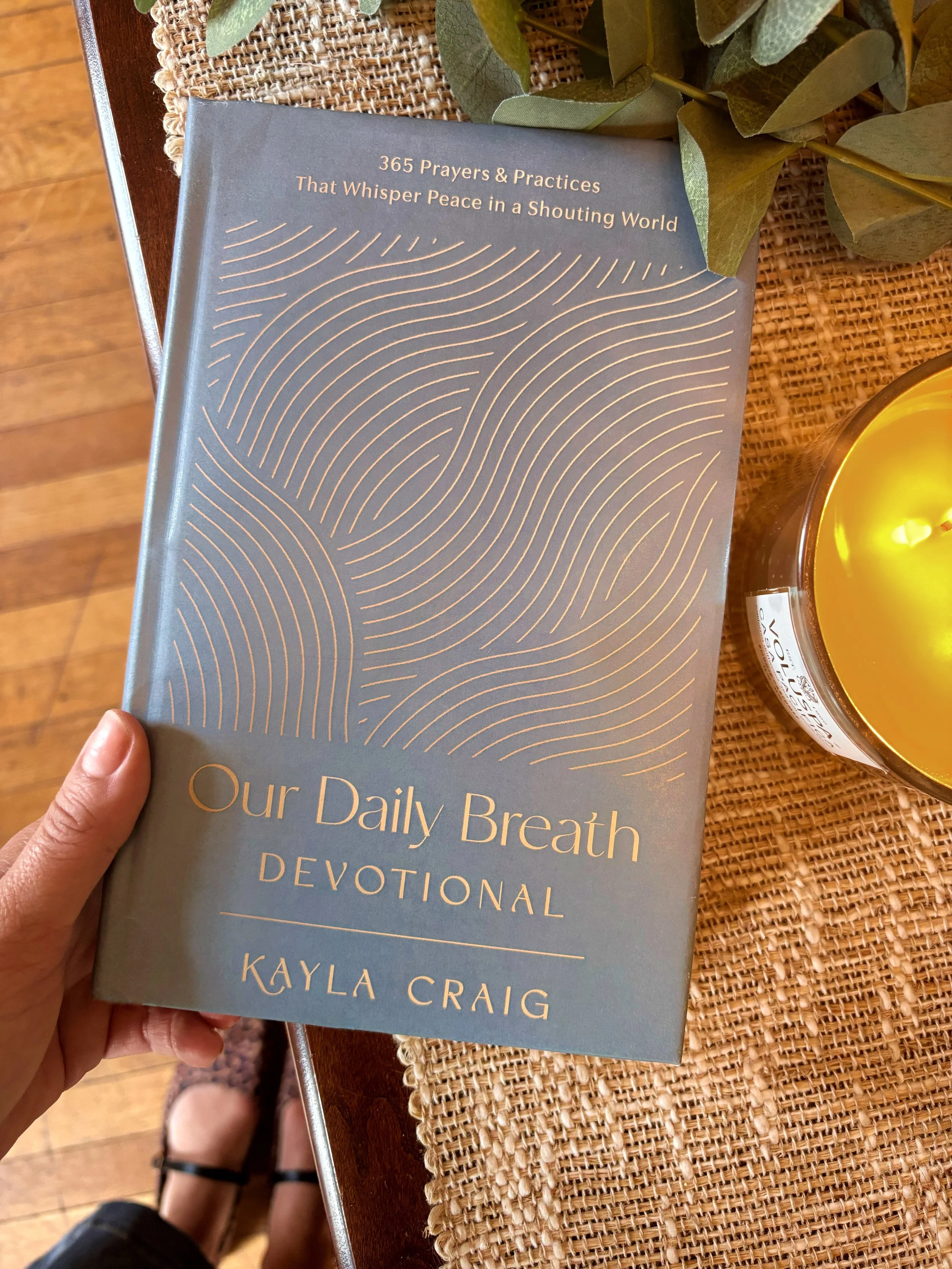Book titled "365 Prayers & Practices That Whisper Peace in a Shouting World" by Kayla Craig, held in a person's hand, on a table with a woven placemat, greenery, and a lit candle.