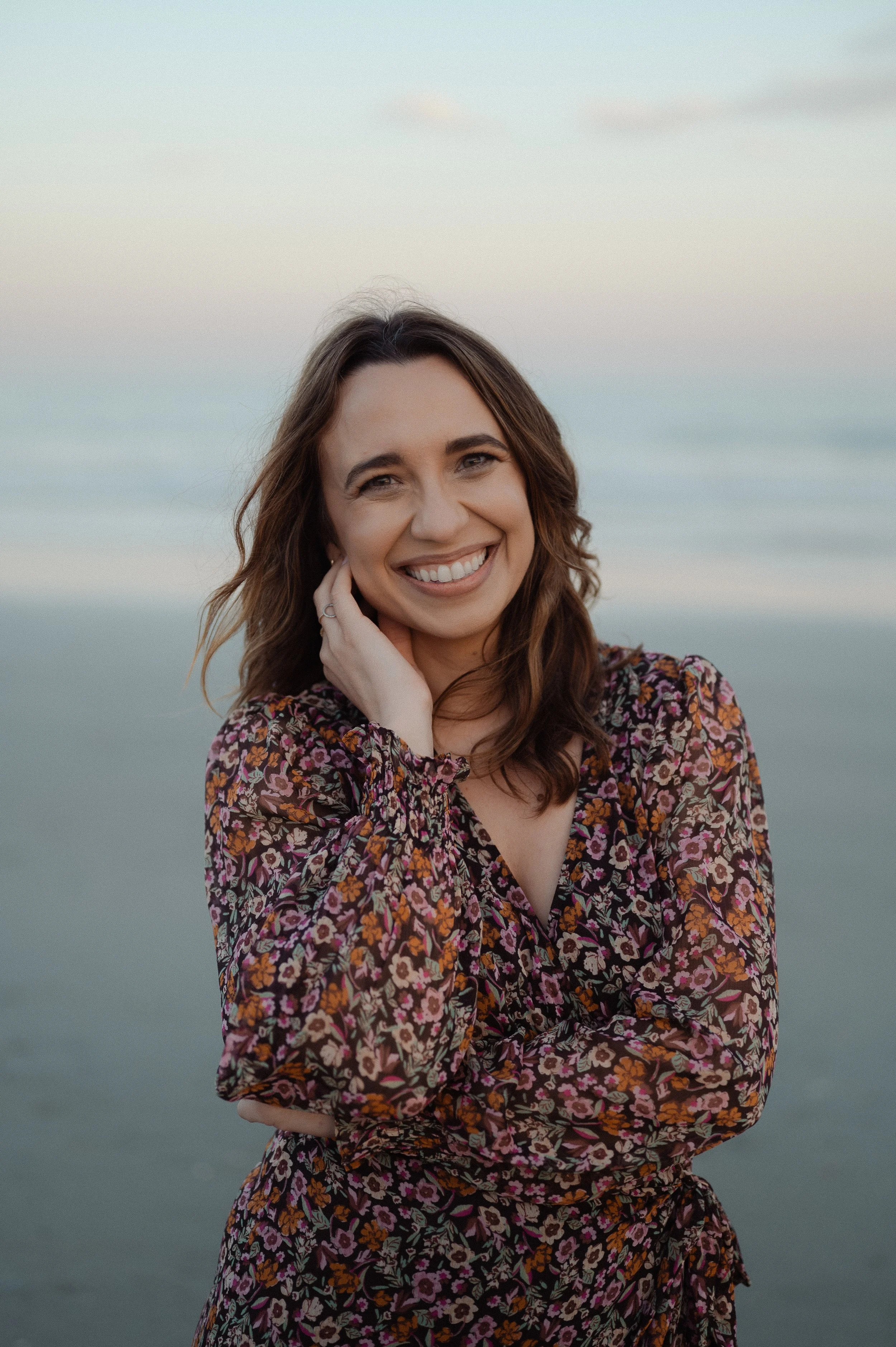 Christian author Kayla Craig with long brown hair wearing a floral dress, smiling with her hand touching her cheek. Background appears to be an outdoor setting with a muted ocean view and a light sky as she honors her new book Our Daily Breath.