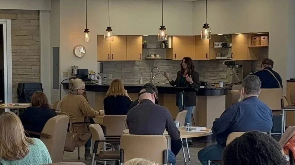 Kayla Craig giving an engaging prayer practices workshop in  a church room with an audience seated at tables, with a kitchen or cafe area in the background.