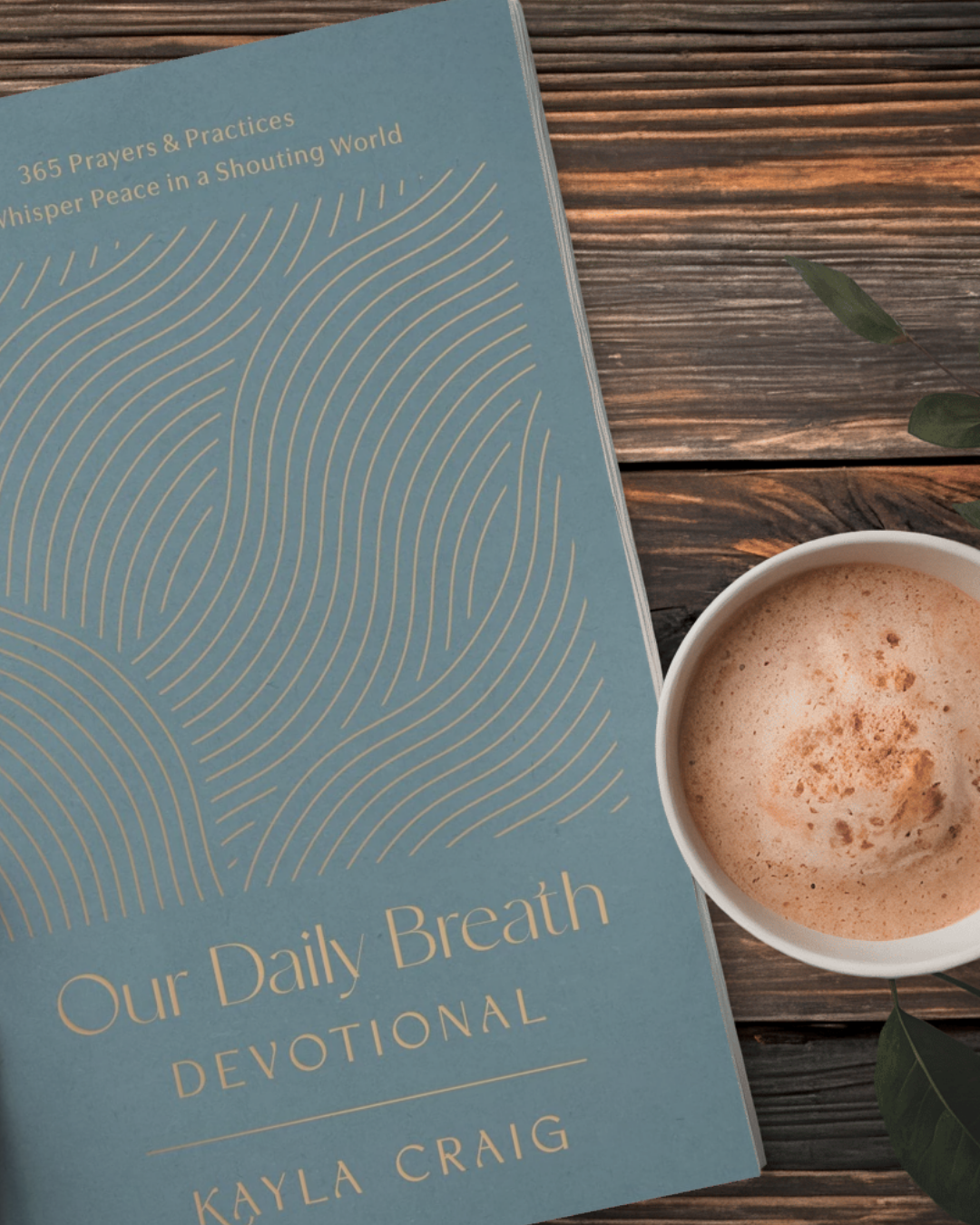 A blue peaceful devotional book titled 'Our Daily Breath: 365 Breath Prayers and Practices that Whisper Peace in a Shouting World' by Kayla Craig lying on a wooden surface next to a white cup of frothy beverage, possibly coffee or hot chocolate.