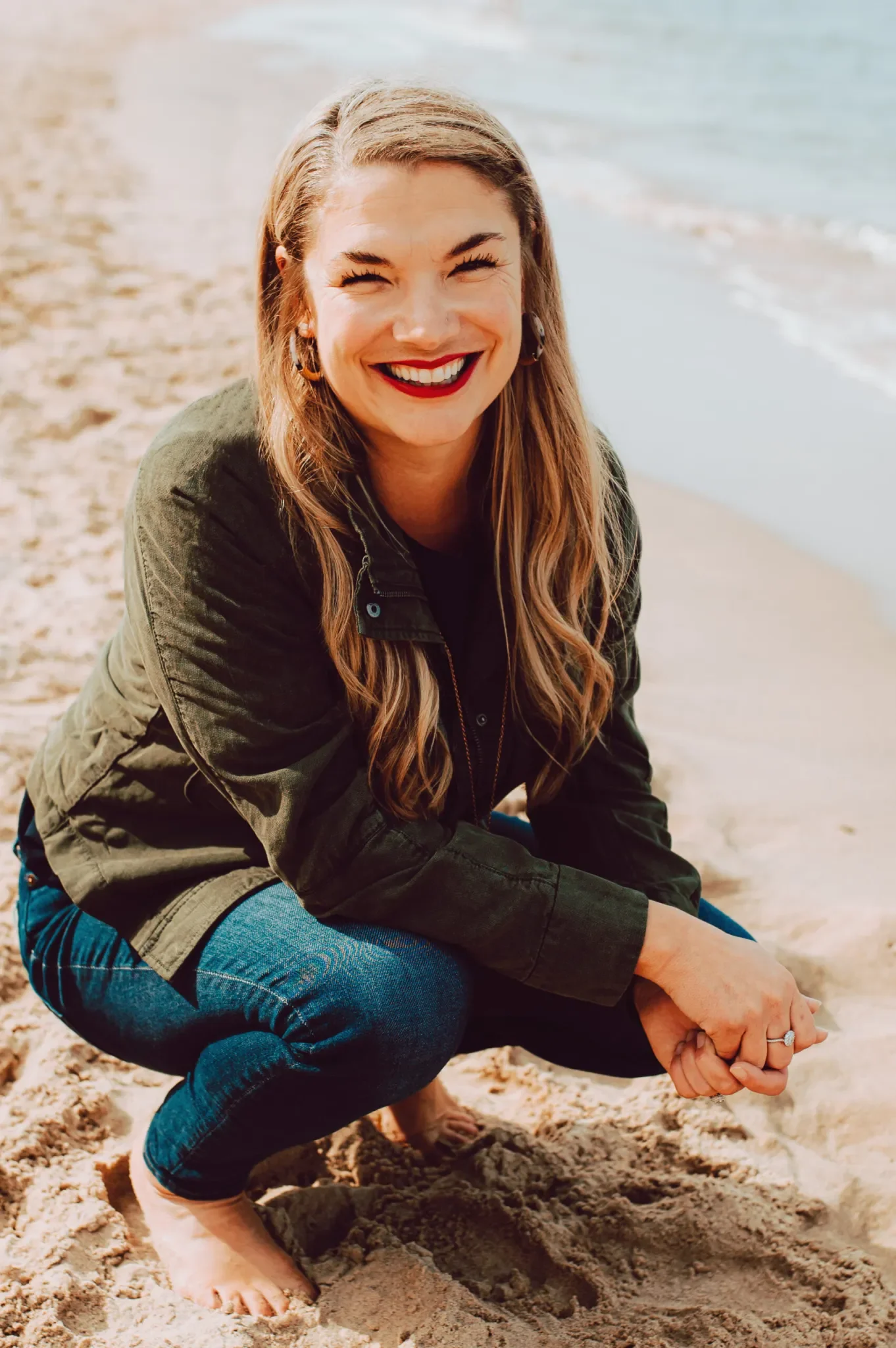 Headshot of bestselling author and therapist Aundi Kolber. Woman with long light brown hair, red lipstick, and earrings smiling on a sandy beach, squatting on her knees with her hands clasped in front of her.