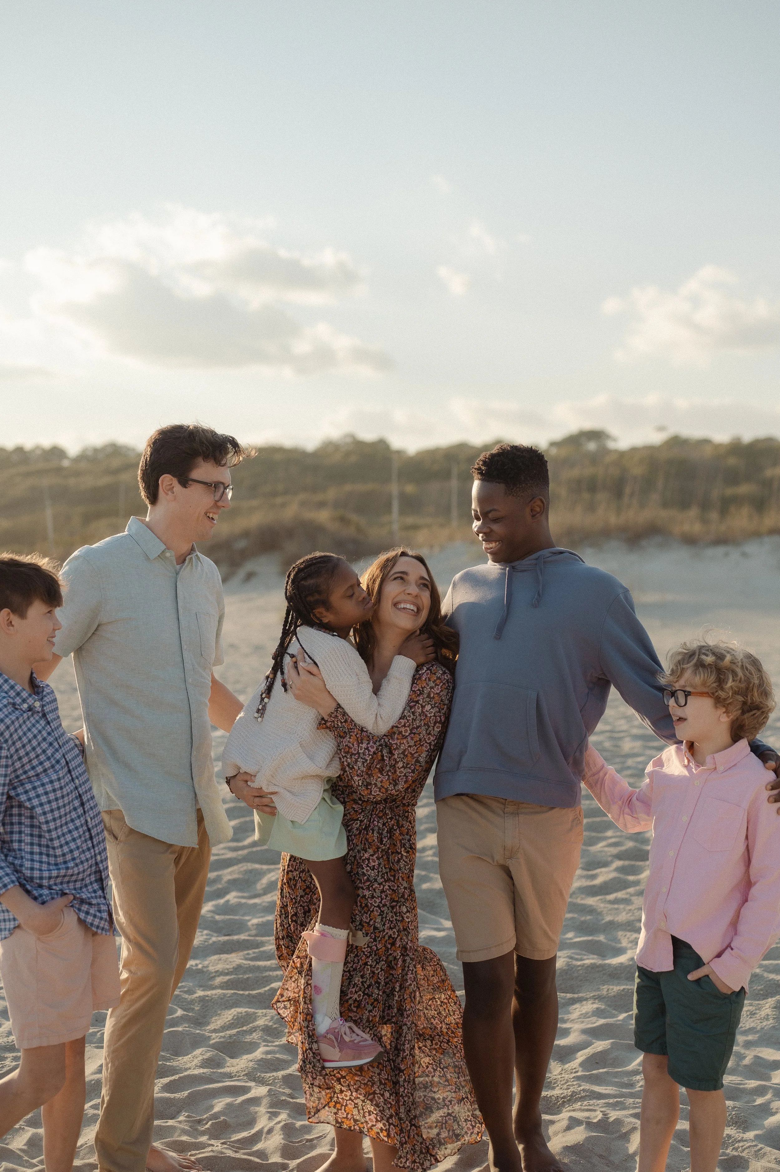 Kayla Craig, author of Our Daily Breath, standing with her family. A diverse group of people on a beach, smiling and enjoying each other's company during sunset.