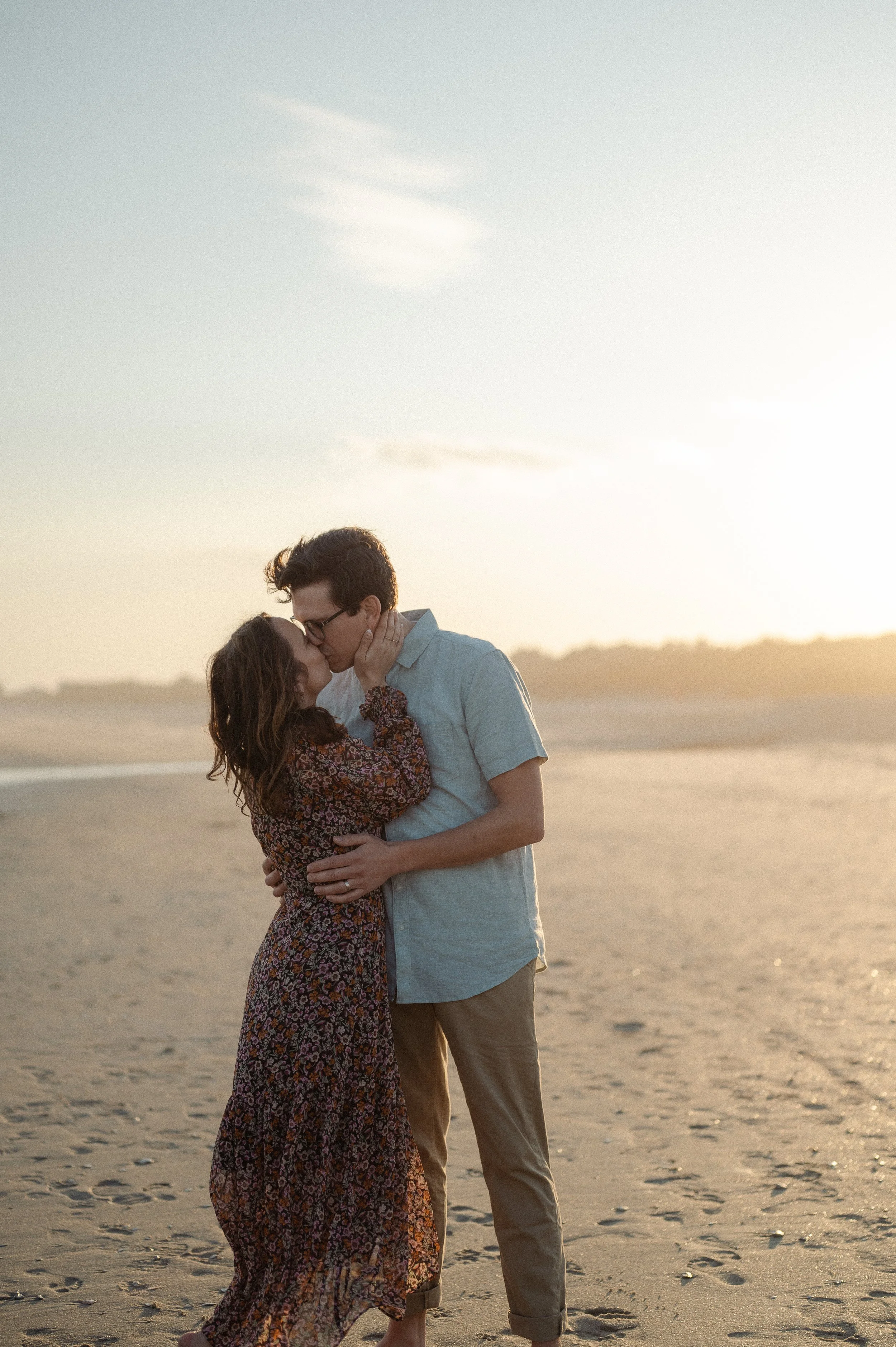 Author Kayla Craig shares a kiss and embrace on a beach at sunset with her husband pastor Jonny Craig. Kayla is wearing a long, patterned dress, and the man is in a short-sleeve button-up shirt and rolled-up pants. The beach is sandy with footprints.
