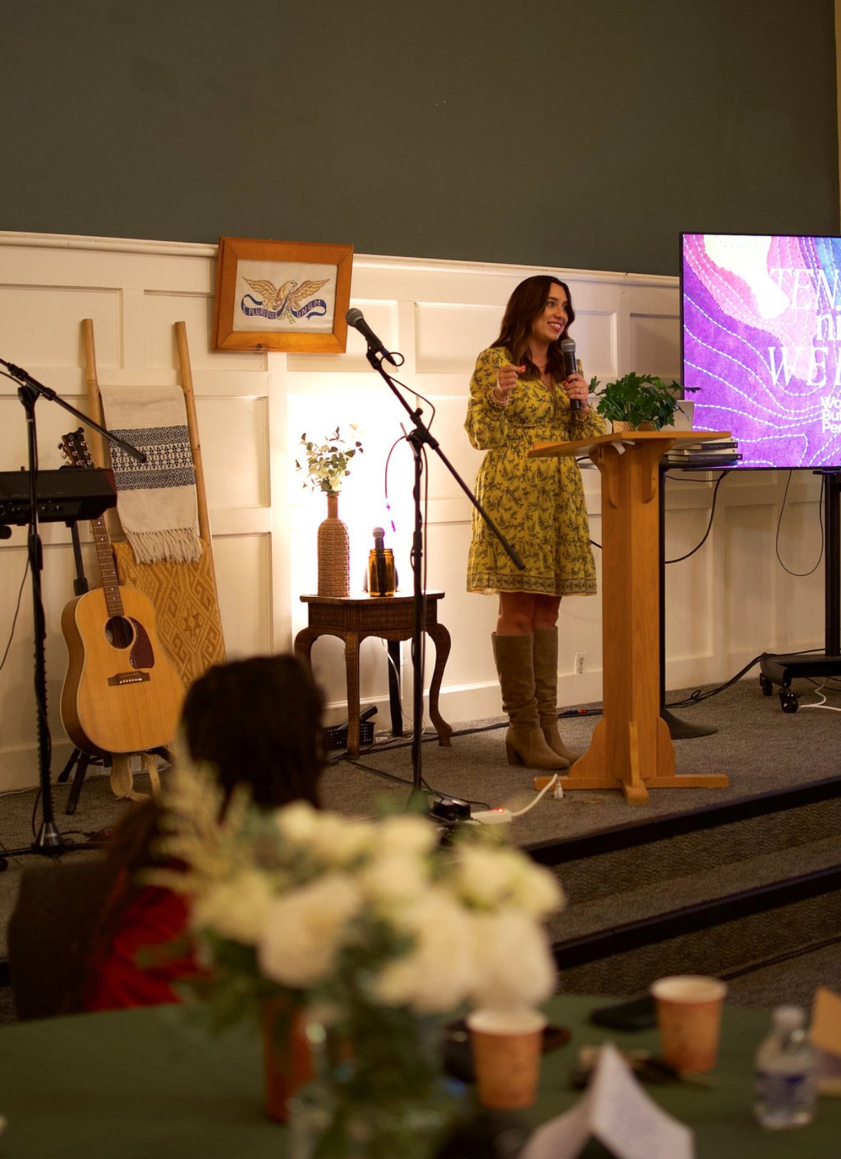 Author Kayla Craig in a yellow floral dress standing at a wooden podium, speaking into a microphone during an indoor event with musical instruments and a large screen behind her.