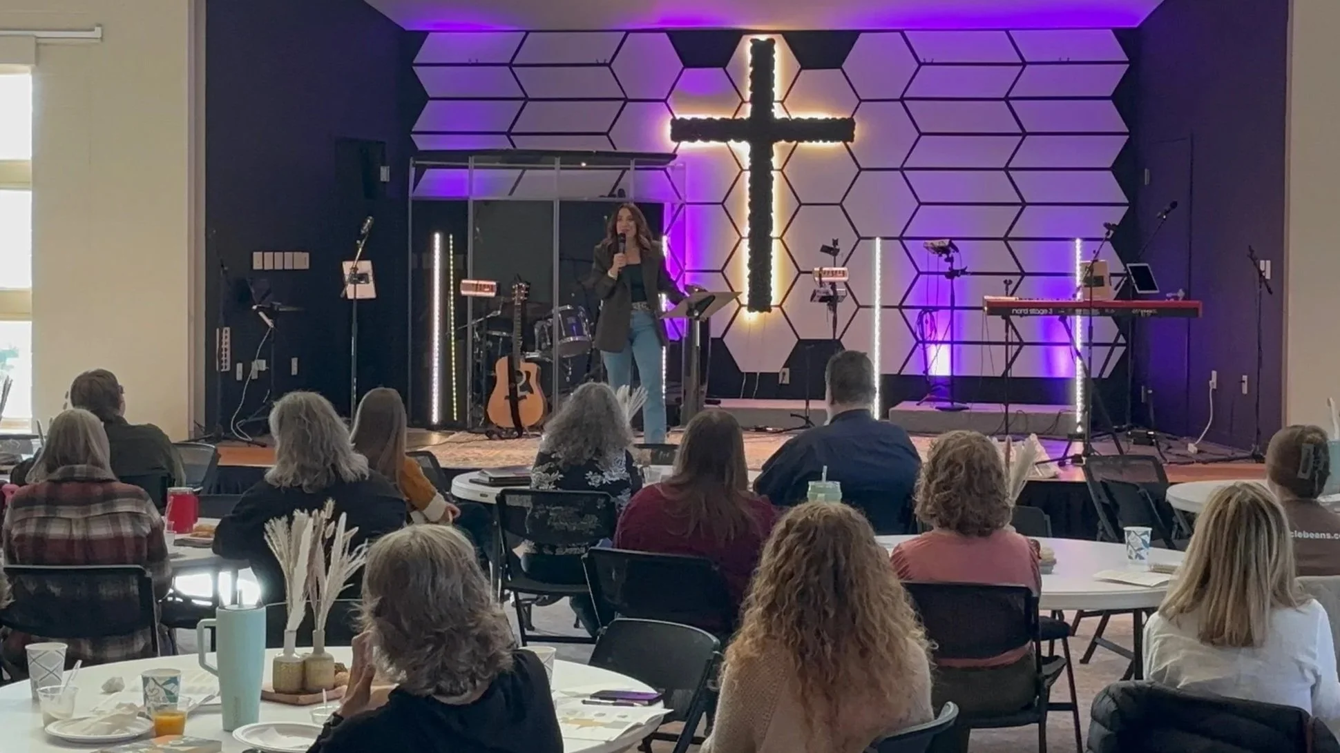 Kayla Craig speaking with a microphone on stage during a large church conference called Woven, with musical instruments behind her and an illuminated cross on the wall. Audience members sit at round tables in a large room.