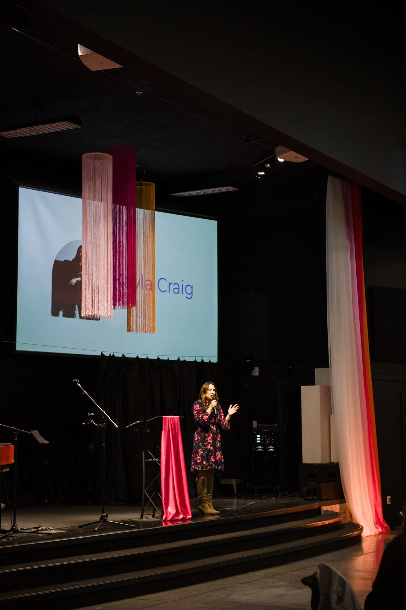 Christian speaker and creator of Liturgies for Parents Kayla Craig standing stage giving a keynote speech or presentation, with a large screen behind her displaying her name and colorful hanging decoration.