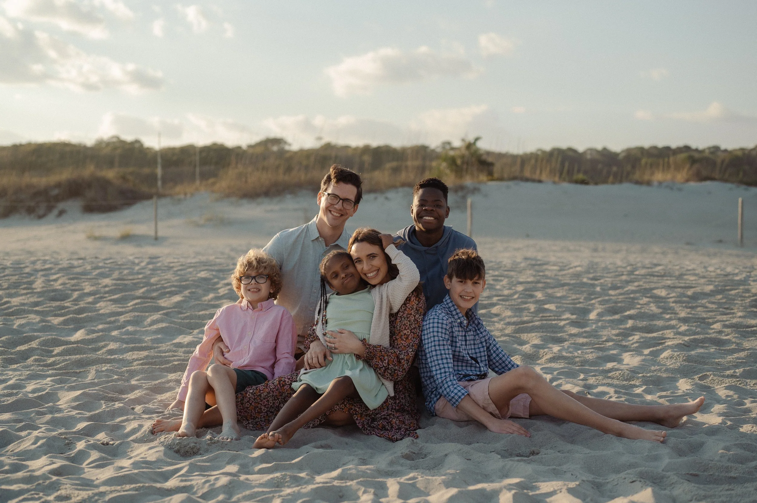 Kayla Craig, author of To Light Their Way, Every Season Sacred, and Our Daily Breath, with a photo of her family, including children and adults, sitting on the sand with a background of dunes and a partly cloudy sky.