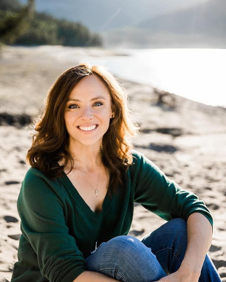 Headshot of bestelling author Sarah Bessey, woman with shoulder-length brown hair smiling while sitting on the beach near the water, wearing a green top and jeans, with the ocean and sky in the background.