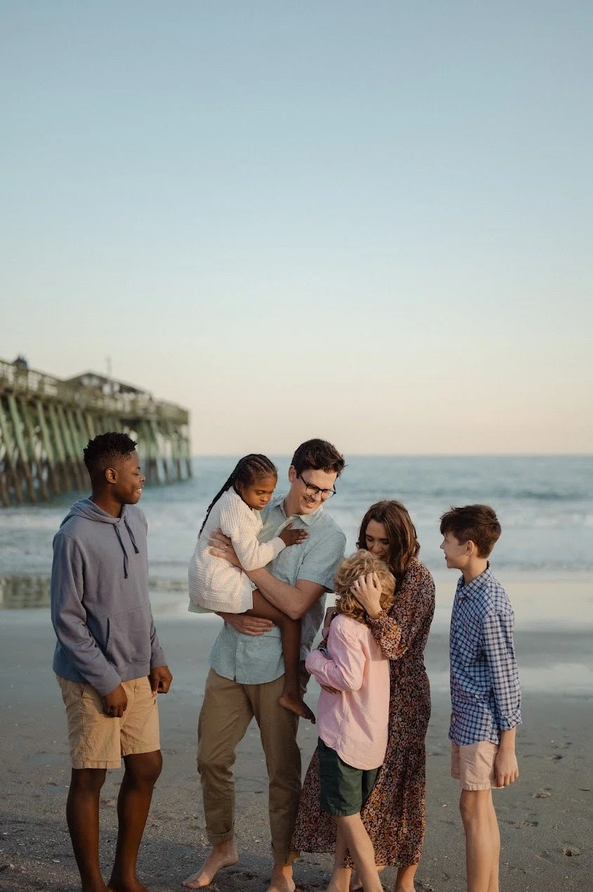 Author Kayla Craig's family, children and adults, enjoying time on a beach with a pier in the background, at sunset, as they celebrate the launch of her newest book Our Daily Breath: 265 Prayers and Practices that Whisper Peace in a Shouting World.