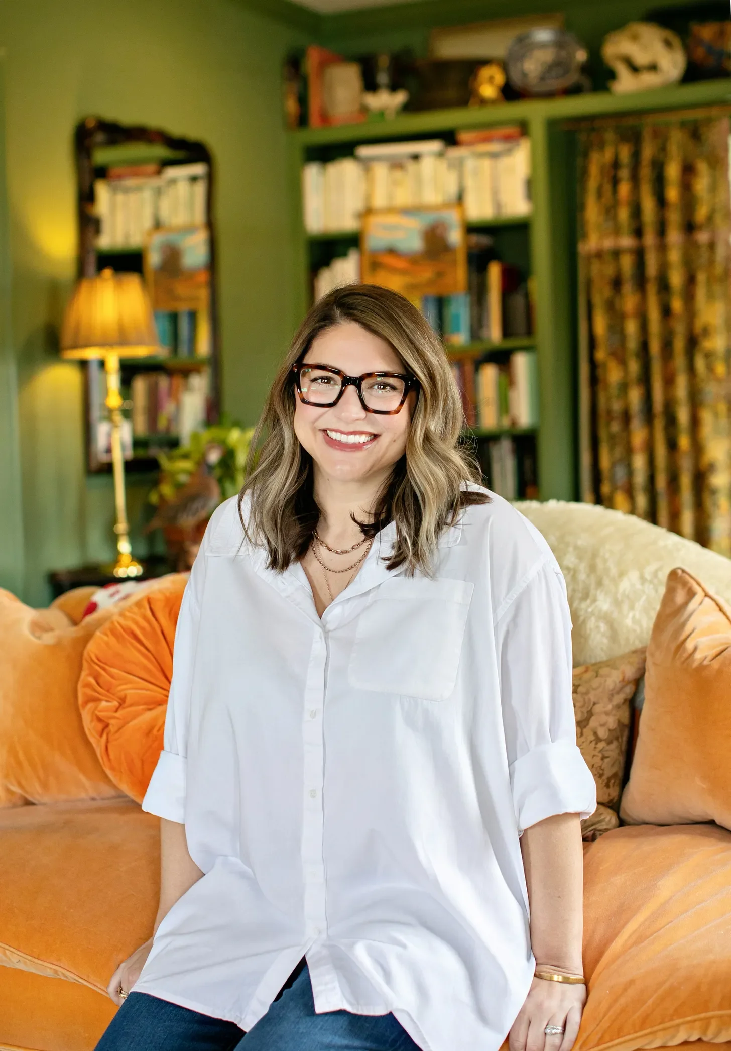 Headshot of Erin H. Moon, author and podcaster.  Woman with shoulder-length brown hair wearing glasses, a white shirt, and jeans, sitting on an orange couch in a room with green walls, bookshelves, curtains, and lamps. She is smiling at the camera.