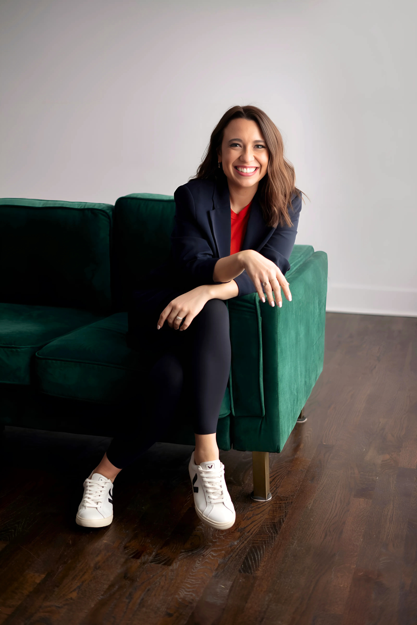 Christian author Kayla Craig is a spiritual director and bestselling author. She is sitting on a green sofa, smiling, wearing a navy blazer, red shirt, black pants, and white sneakers, with a plain white wall in the background.