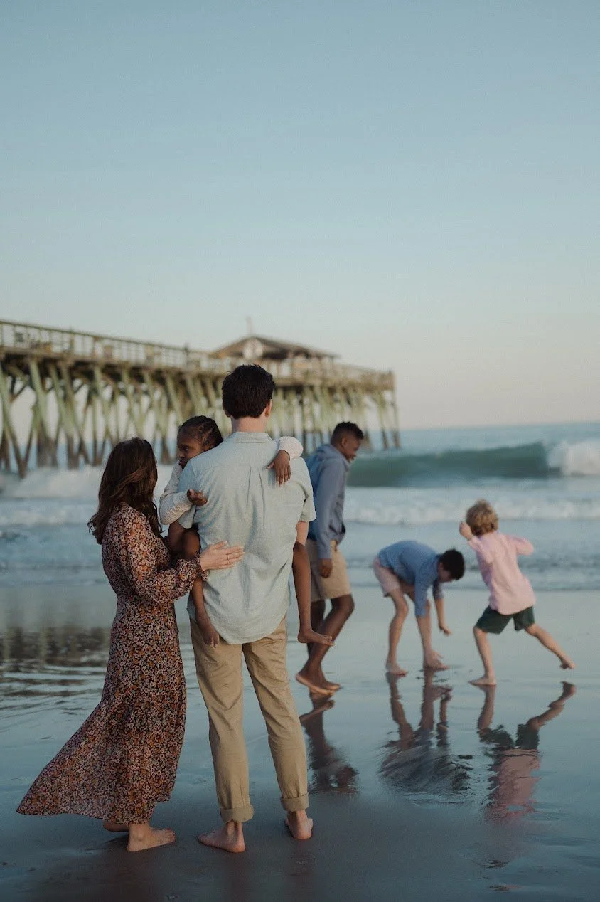 Author Kayla Craig's family. A family of six enjoying time at the beach near a pier, with some children playing near the water and an adult carrying a child.