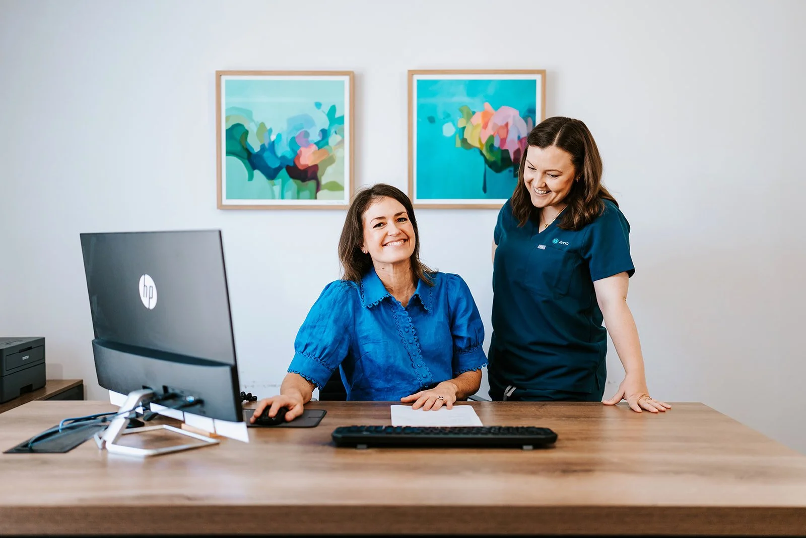 Dr Emma Tetstall and Midwife Anna, one sitting at a desk and one standing next to her, smiling. The woman at the desk is using a computer mouse, with abstract art pieces hanging on the wall behind them.