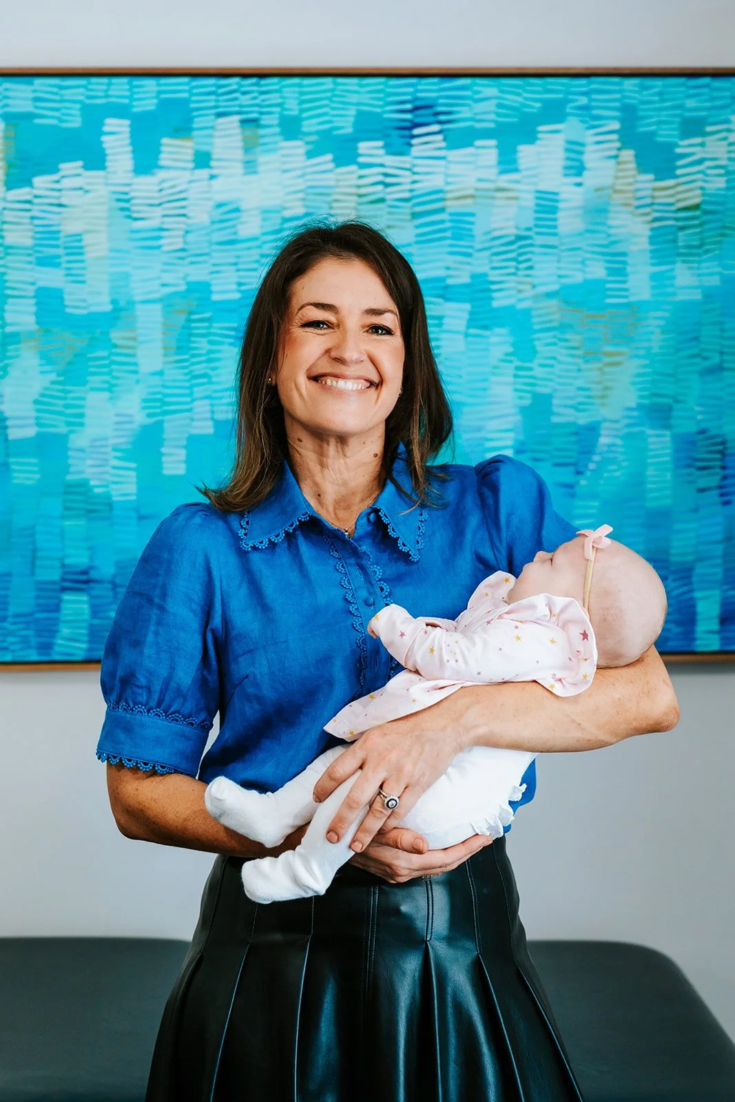 Dr Emma Tetstall wearing a blue blouse, smiling and holding a baby with no hair, dressed in light-colored pajamas, against a blue patterned background.