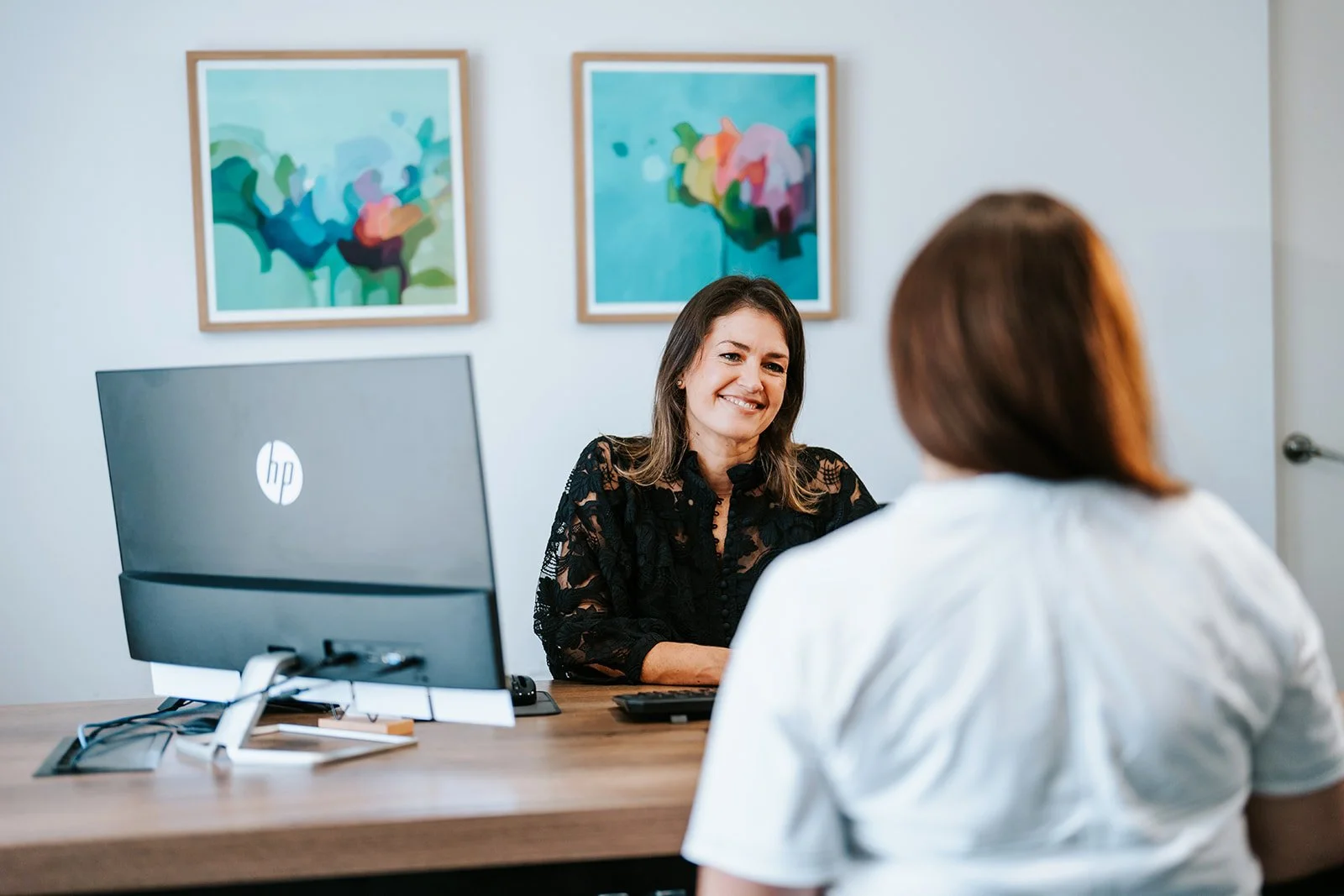 Dr Emma Tetstall sitting at a desk smiling and talking to another woman with her back to the camera. There is a computer monitor and abstract paintings on the wall.