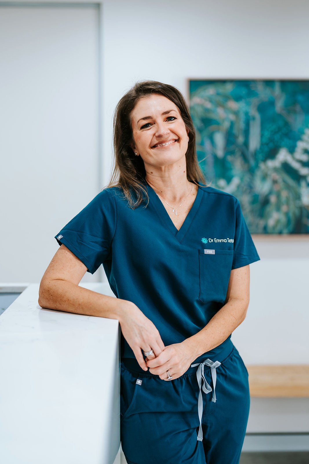 Dr Emma Tetstall wearing blue medical scrubs standing in a clinic or hospital setting, smiling, with a painting on the wall behind her.