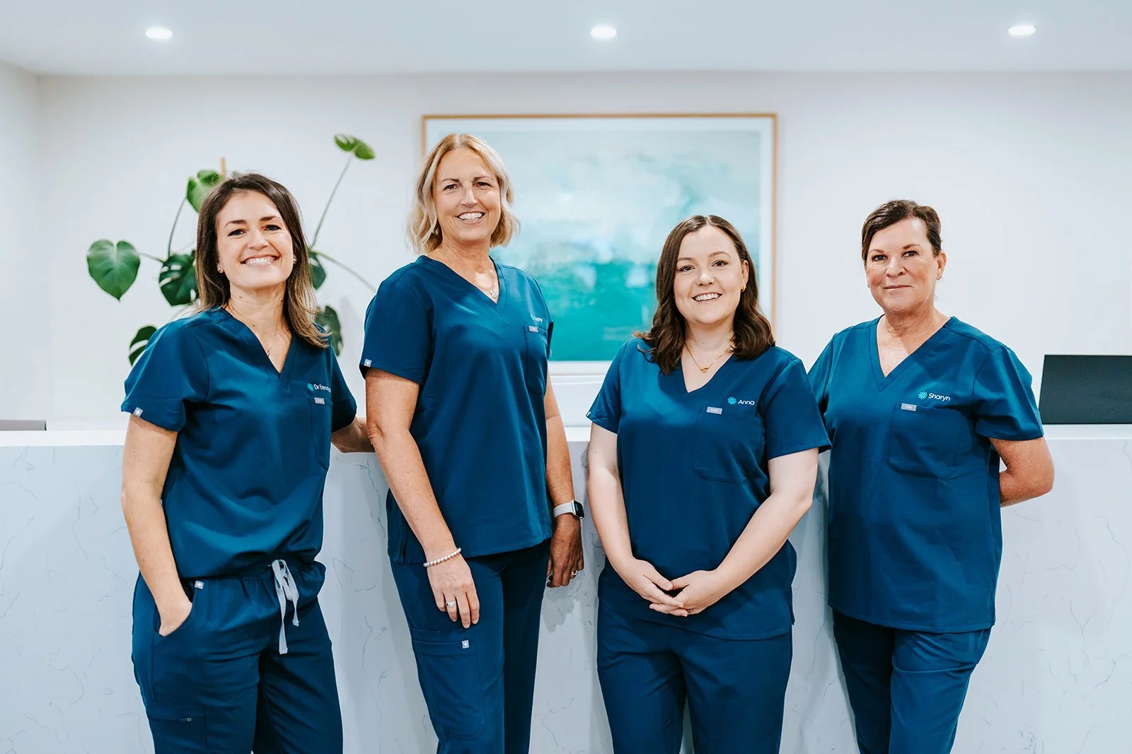 Four women in blue medical scrubs standing behind a white marble reception desk in a healthcare setting, with a plant and framed artwork on the wall behind them.