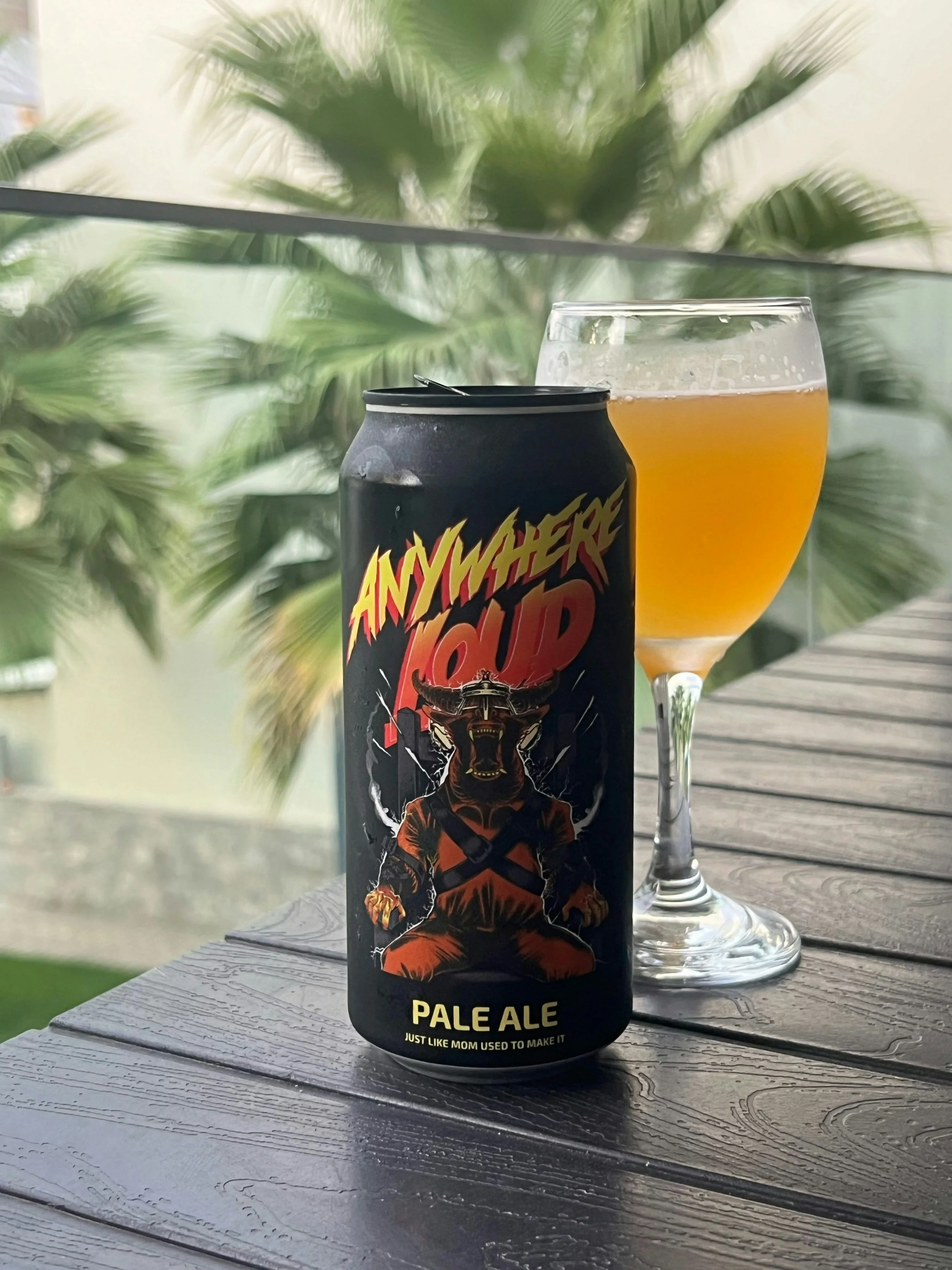 A can of Anywher Mud Pale Ale beer and a glass of beer on a wooden table with palm trees in the background.